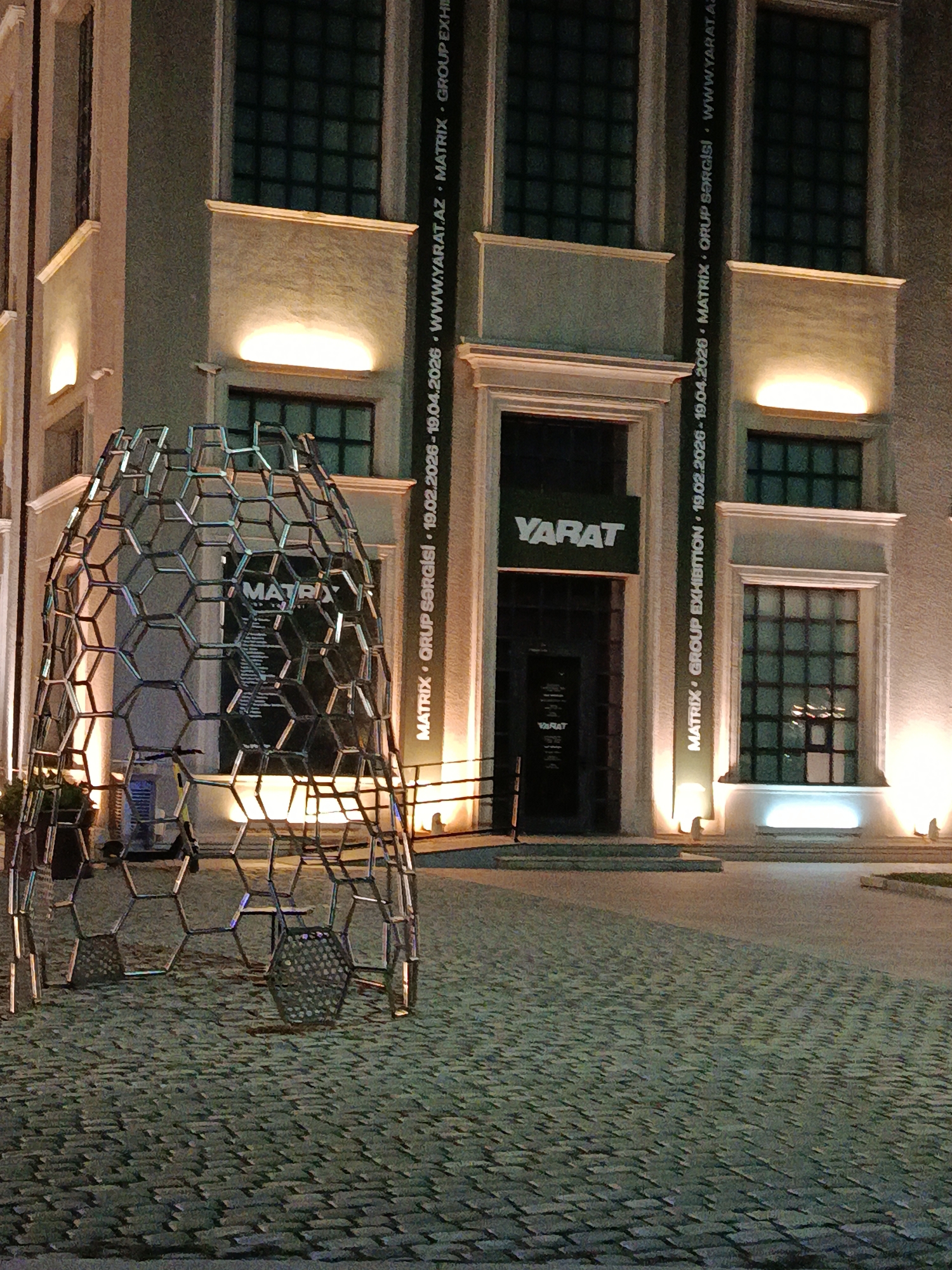 The image depicts the exterior of the YARAT Contemporary Art Space in Sabail Raion, Azerbaijan, at night. In the foreground, a large, geodesic dome-like sculpture constructed from interconnected hexagonal metal frames is positioned on a cobblestone plaza. The sculpture is lit from below, casting a subtle glow. Behind the sculpture, the facade of the YARAT building is visible. The building features large, dark-paned windows and is illuminated by warm, upward-facing lights. A prominent vertical banner on the building's facade displays text in white, reading "MATRIX GROUP EXHIBITION 19.02.2026 - 19.04.2026 WWW.YARAT.AZ MATRIX GROUP EXHIBITION". The entrance to the building, marked with a "YARAT" sign, is also visible. The plaza is paved with light-colored cobblestones arranged in a regular pattern. The overall scene suggests an outdoor exhibition or event space, with the sculpture serving as a focal point.