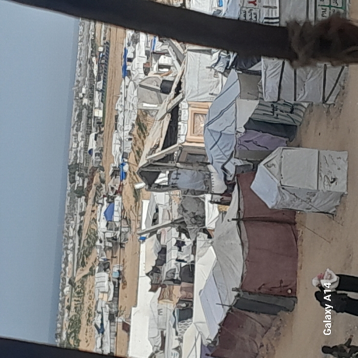 A densely populated refugee camp stretches across a sandy landscape, filled with makeshift tents and shelters. The tents, varying in color and construction, appear crowded together, suggesting a limited space. In the foreground, structures seem more established, possibly indicating longer habitation. The overall view suggests challenging living conditions and a desperate need for space and resources.