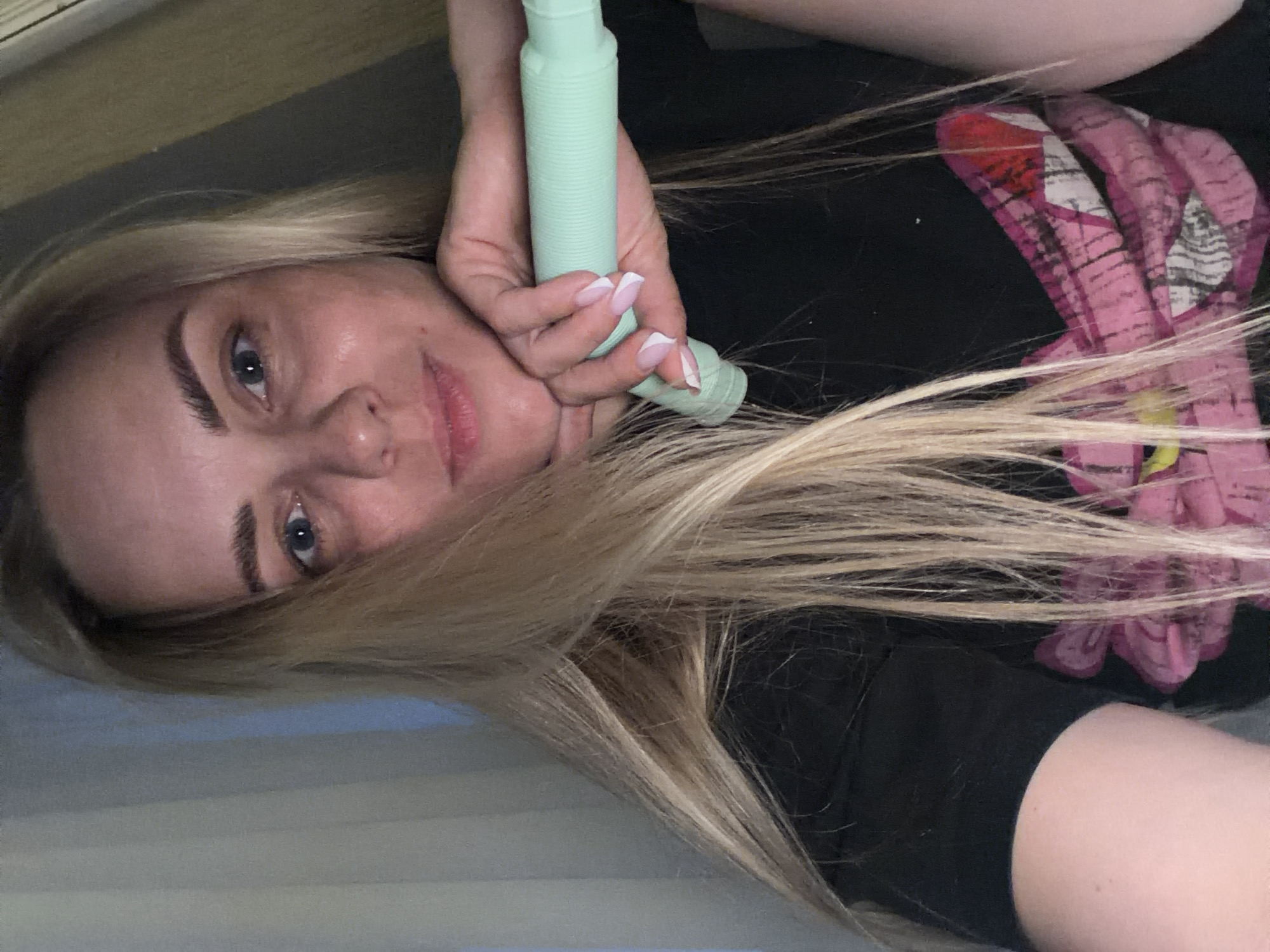 A blonde woman is lying down, holding a light green, corrugated plastic tube. She is wearing a black shirt with a pink graphic. The background is a neutral beige and gray. There is no indication of the city of Votkinsk, Russia.