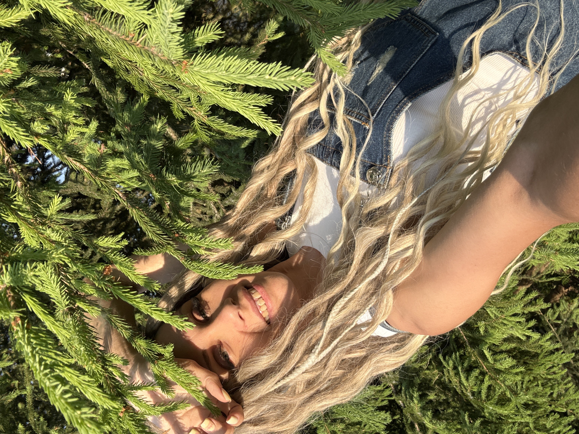 The image shows a close-up, upside-down, outdoor shot of a woman lying among pine branches. She has long, blonde, braided hair that is partially covering her face. Her eyes are looking towards the camera, and she is smiling with her teeth showing. She is wearing a white top and a denim vest. The pine branches are a vibrant green, and the lighting suggests it is a sunny day. The setting appears to be a forest or wooded area, and the location context is Rusanovo, Russia. The overall emotion conveyed is one of joy and relaxation. There is no visible text in the image.
