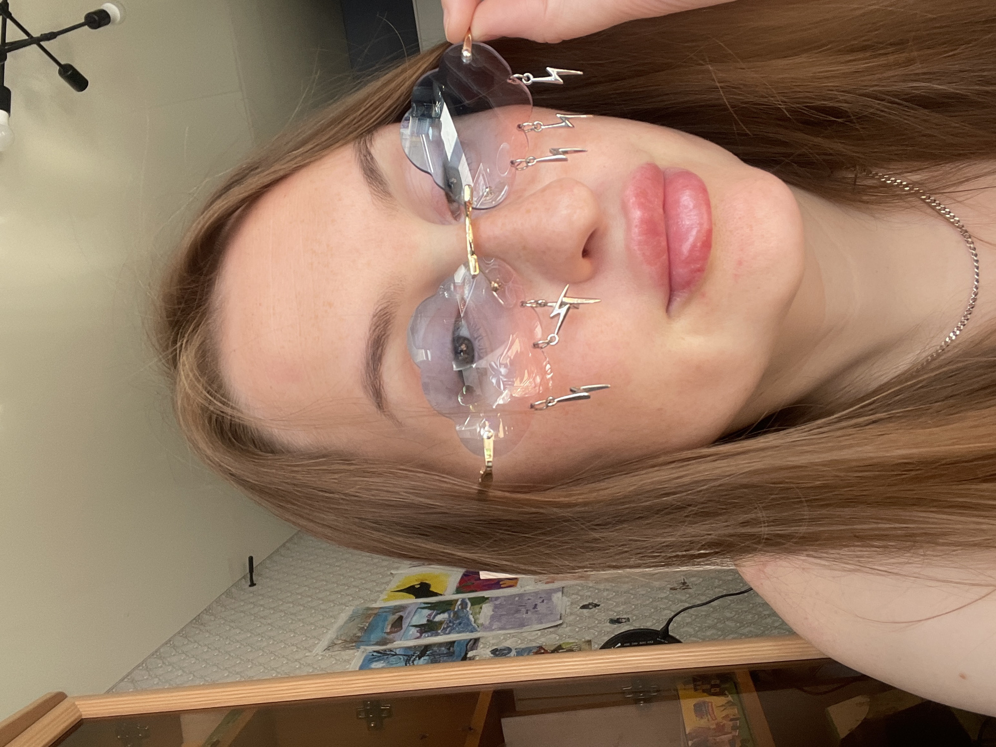 A young woman with long, light brown hair and freckles is posing for a photo indoors. She is wearing novelty cloud-shaped sunglasses with lightning bolt charms dangling from them. She is looking at the camera and her right hand is adjusting the sunglasses. The background shows a wall with wallpaper, some framed pictures, and a cabinet. The scene appears to be a bedroom or living area. The location is specified as Syurkovo, Russia.