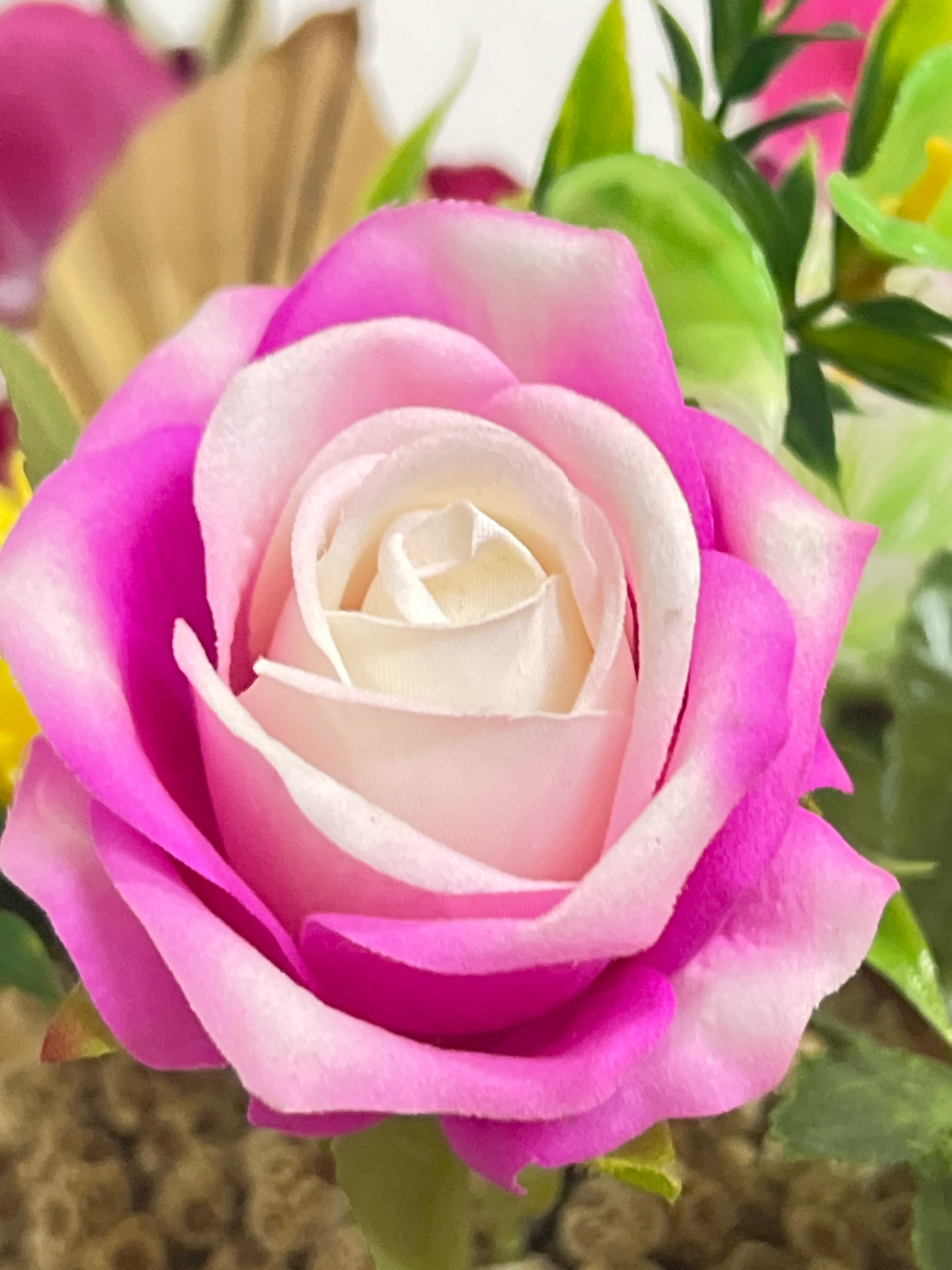 The image is a close-up shot of a vibrant pink and white rose, likely artificial, with soft lighting. The rose is the primary subject, with its petals unfurling in a spiral pattern. The outer petals are a rich pink, fading to a lighter pink and then white towards the center. The background is a blur of greens and other muted colors, suggesting other floral elements or foliage. The overall impression is one of delicate beauty and detailed texture. There is no visible indication of any specific location in Denpasar, Indonesia.