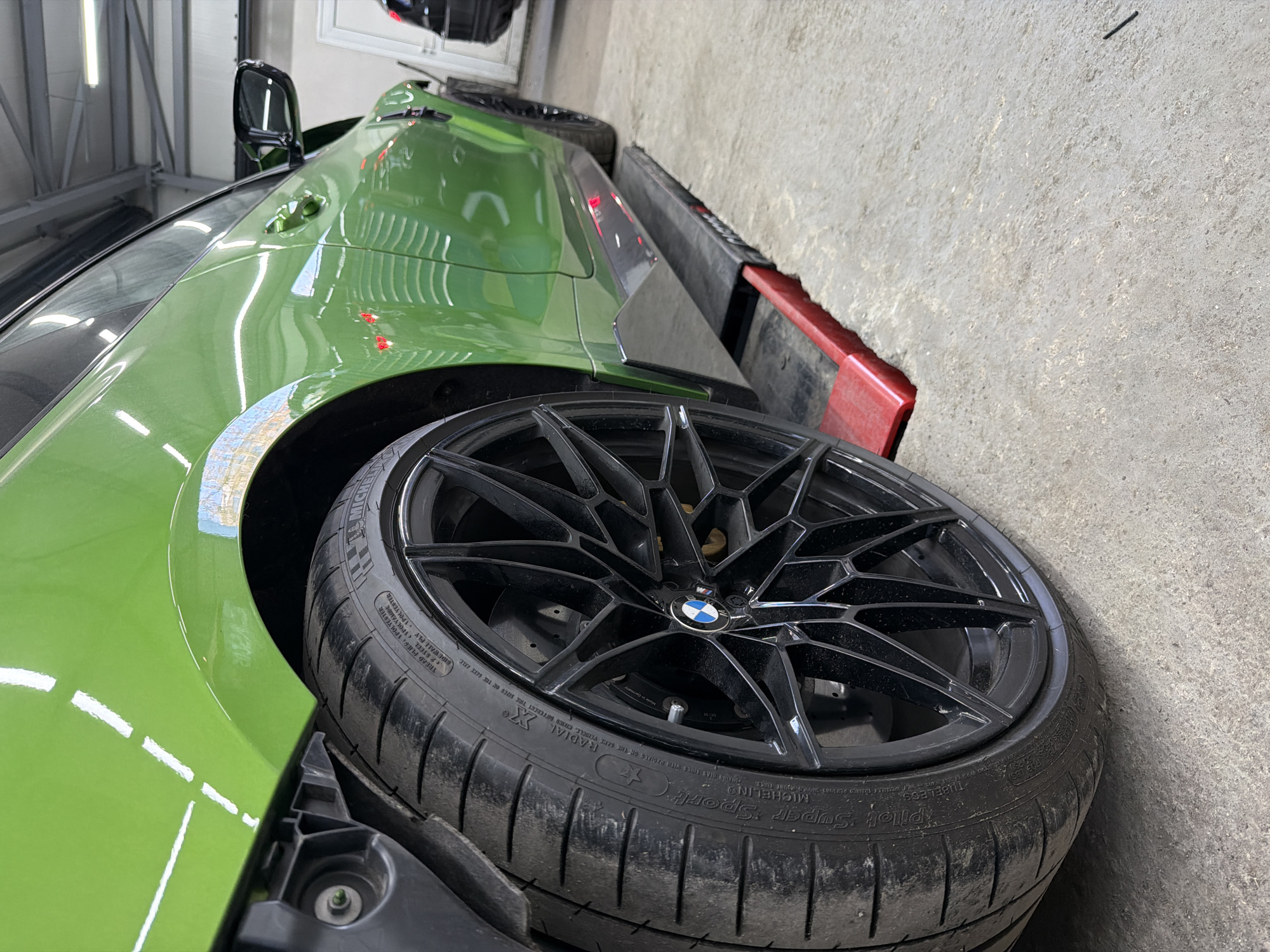 The tilted image captures a close-up of a vibrant green car, likely a BMW due to the visible logo on the wheel, in an indoor setting that resembles a garage or workshop. The car's glossy, reflective paint job shows clear reflections of overhead lighting. The primary focus is on the car's front wheel, which is black with a distinctive multi-spoke design. The tire mounted on the wheel is a Michelin Pilot Super Sport, with the brand and model name clearly legible as "MICHELIN" and "Pilot Super Sport," along with "TUBELESS" and "RADIAL X." Other visible text on the tire includes "TREAD PLIES POLYESTER" and "STEEL POLYAMIDE." The car is positioned next to a low red and black barrier or curb. The background features a light gray concrete floor and wall, and some metallic structures are partially visible. The scene suggests the car is stationary, possibly undergoing detailing, maintenance, or storage.