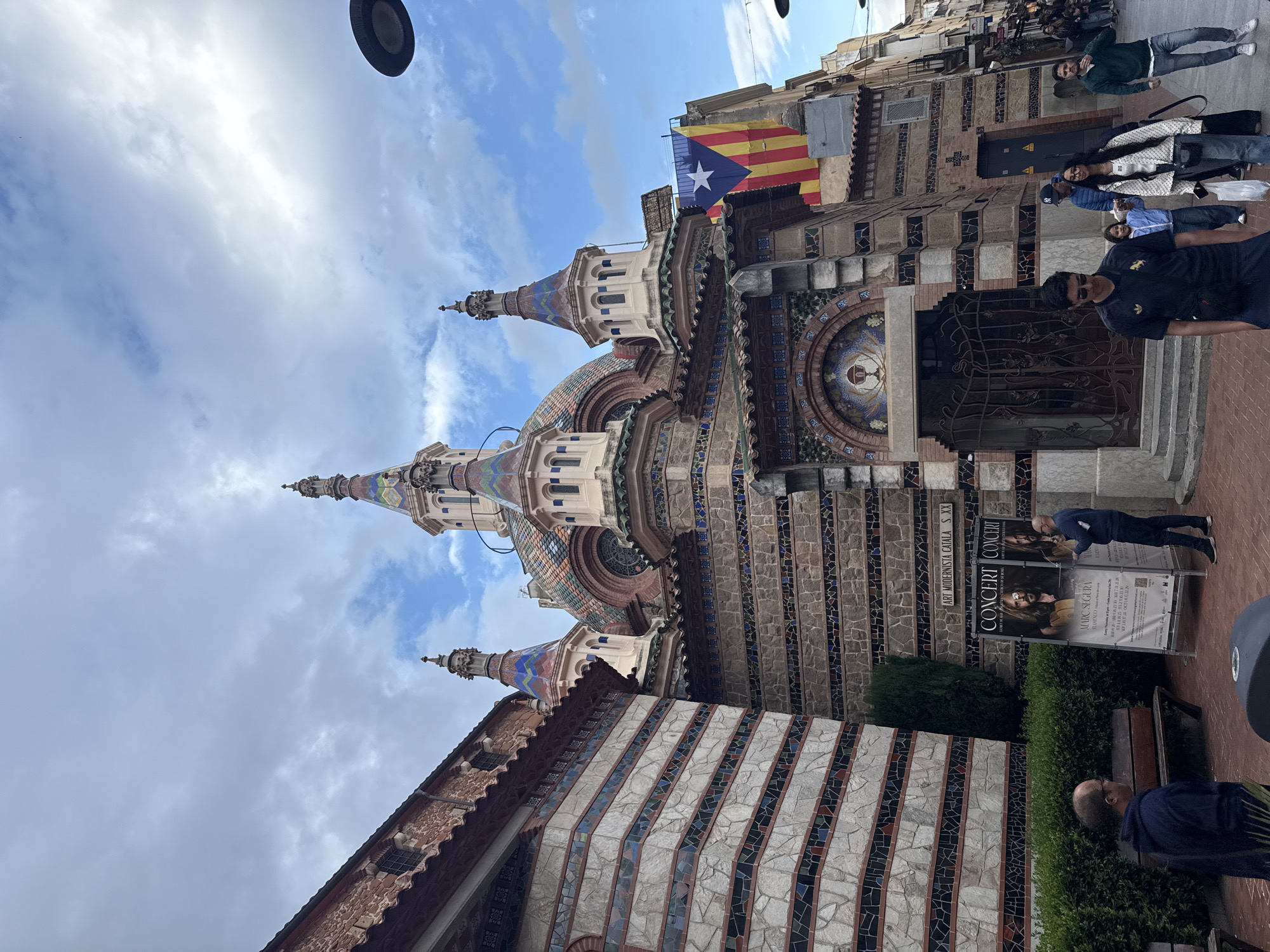 Here is a detailed description of the image:
**Overview:** The image is a vertical shot of the Sant Romà Church in Lloret de Mar, Spain. The church's unique modernist architecture, colorful tiled spires, and mosaic-decorated facade are prominently featured.
**Architecture:** The church shows a blend of architectural styles, with a predominant modernist design characterized by colorful tiles and irregular shapes. The mosaic work and the spires give it a distinctive appearance. The facade has a large mosaic depicting a chalice with people in front of it.
**People:** People are seen in front of the church.
**Objects:**
* Catalan flag hanging.
* Concert posters.
* A bench.
* Street lamps.
**Actions:** People are interacting with the church, standing and observing.
**Environment:** The setting is a street in Lloret de Mar, with the church facing the street. Other buildings are visible in the background, suggesting a dense urban environment. The sky is partly cloudy.