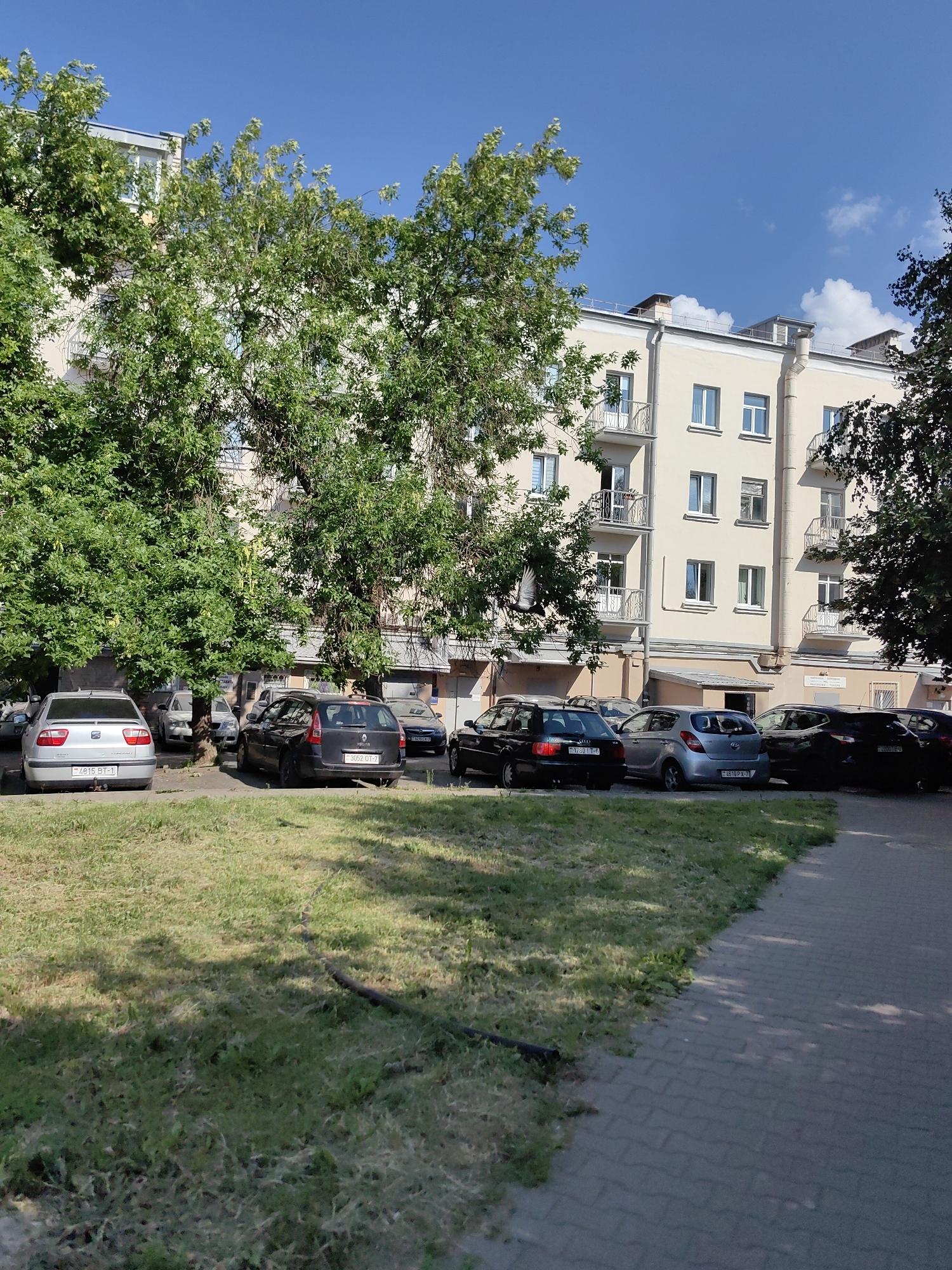This is a daylight, eye-level, medium shot of a parking lot adjacent to a multi-story apartment building in Minsk, Belarus. A light yellow apartment building with multiple windows and balconies dominates the background. In the foreground, a strip of green grass is bordered by a paved walkway with cobblestone-like patterns. Several cars are parked in a line between the grass and the building. The cars include a silver sedan, a black station wagon, a light blue hatchback, and several other dark-colored vehicles. The sky is clear and blue with a few scattered white clouds. The scene appears to be a typical residential area.