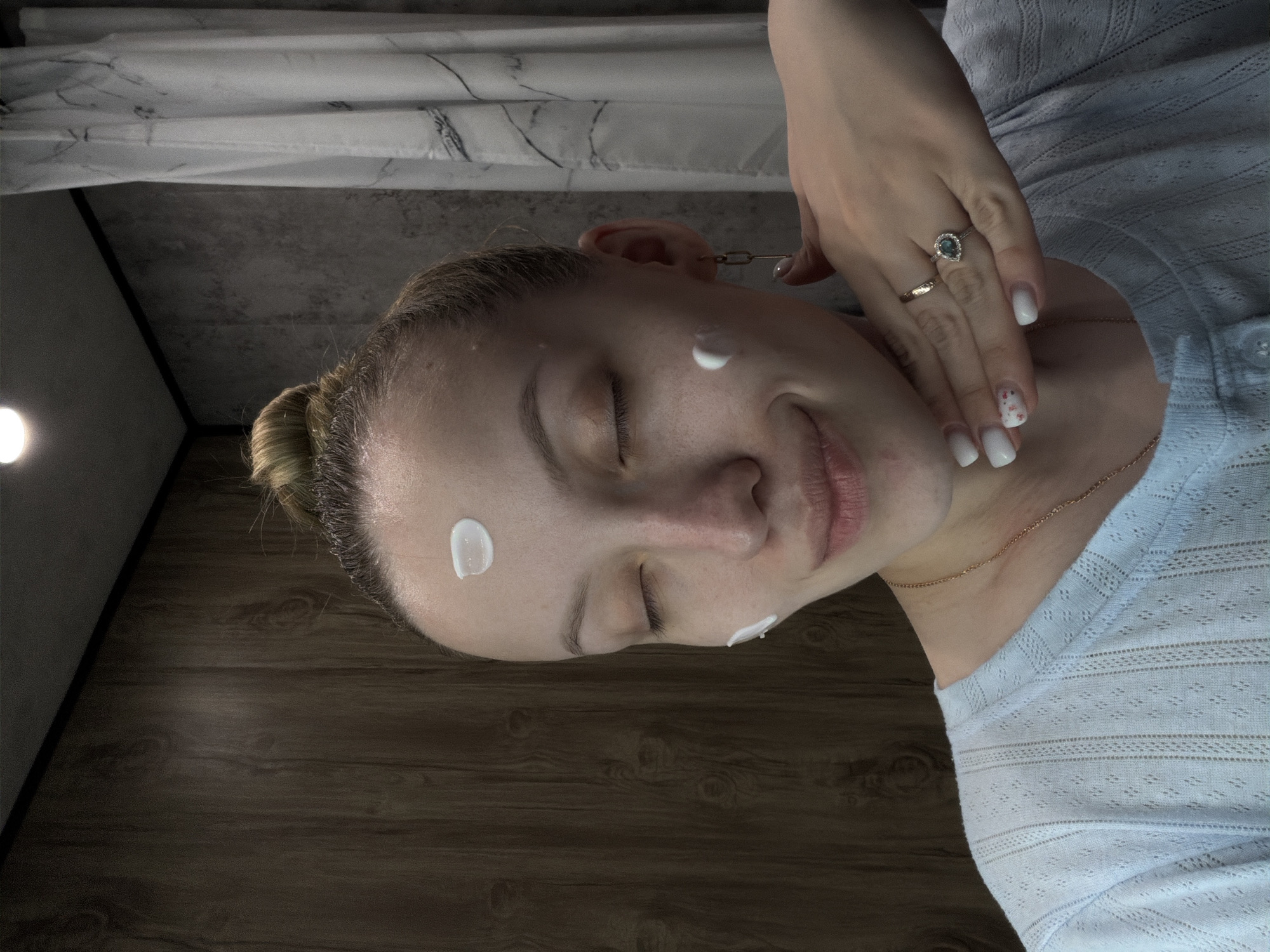 A young woman with her hair in a bun is shown with dots of white cream on her forehead and cheek. Her eyes are closed, and she is smiling slightly. She is wearing a light blue top, a delicate gold necklace, and two rings on her right hand. Her fingernails are painted white, with one nail featuring a delicate floral design. The background includes a wood-patterned surface, a grey wall, and a white curtain or sheet with a marble-like pattern. There is no information suggesting the location of Reshetnikovo, Russia, is specifically represented in the image.