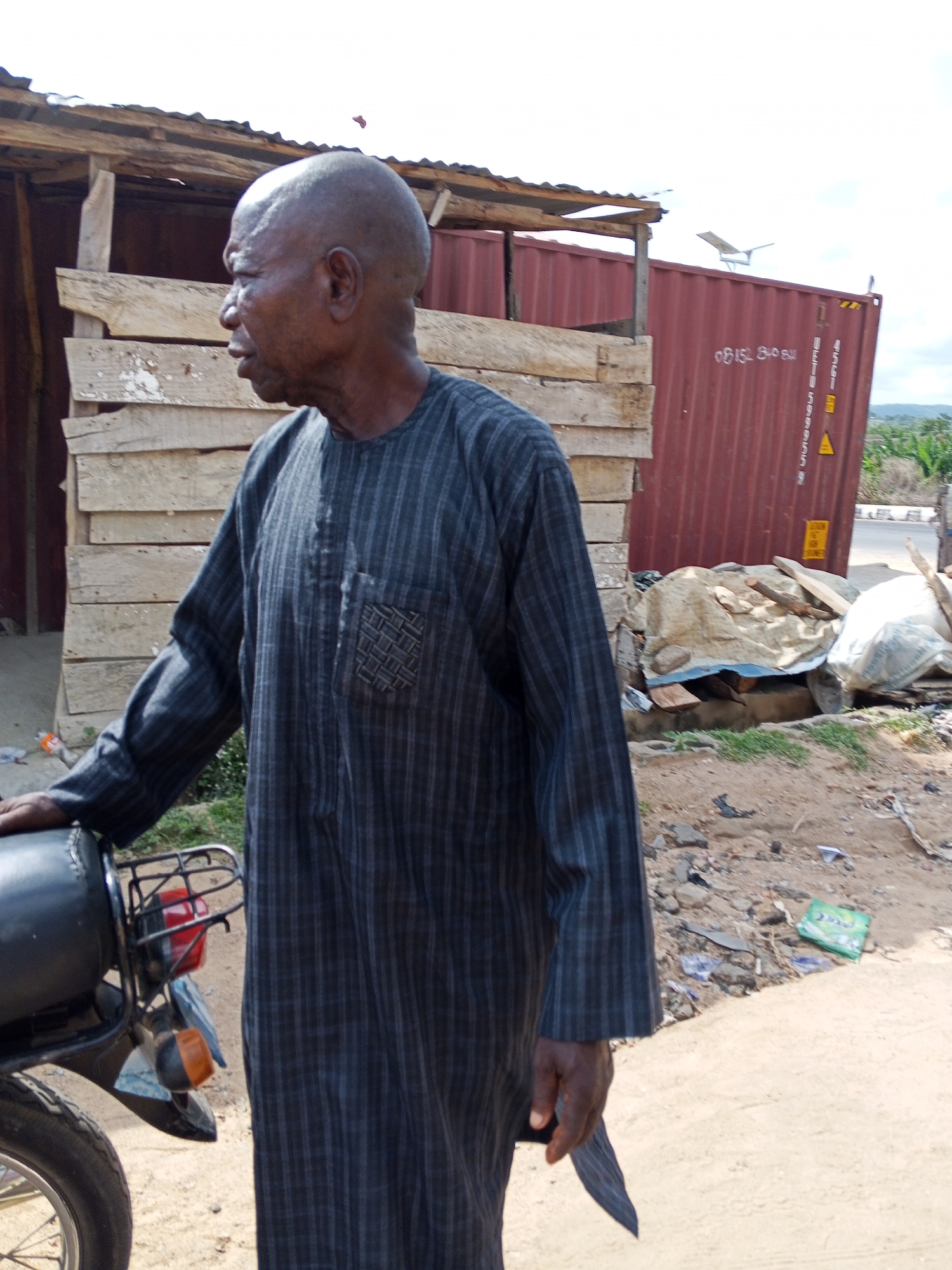 The image depicts an elderly, bald-headed man in a striped, long-sleeved garment standing next to a motorcycle. He is looking to the left. In the background, there's a dilapidated wooden structure, a red shipping container with faded markings, and general debris scattered on the ground. A solar-powered street light stands atop the shipping container. The location is identifiable as Zango-ndaji, Nigeria based on the presence of the specific type of building, the clothing of the man, and the general environment.
