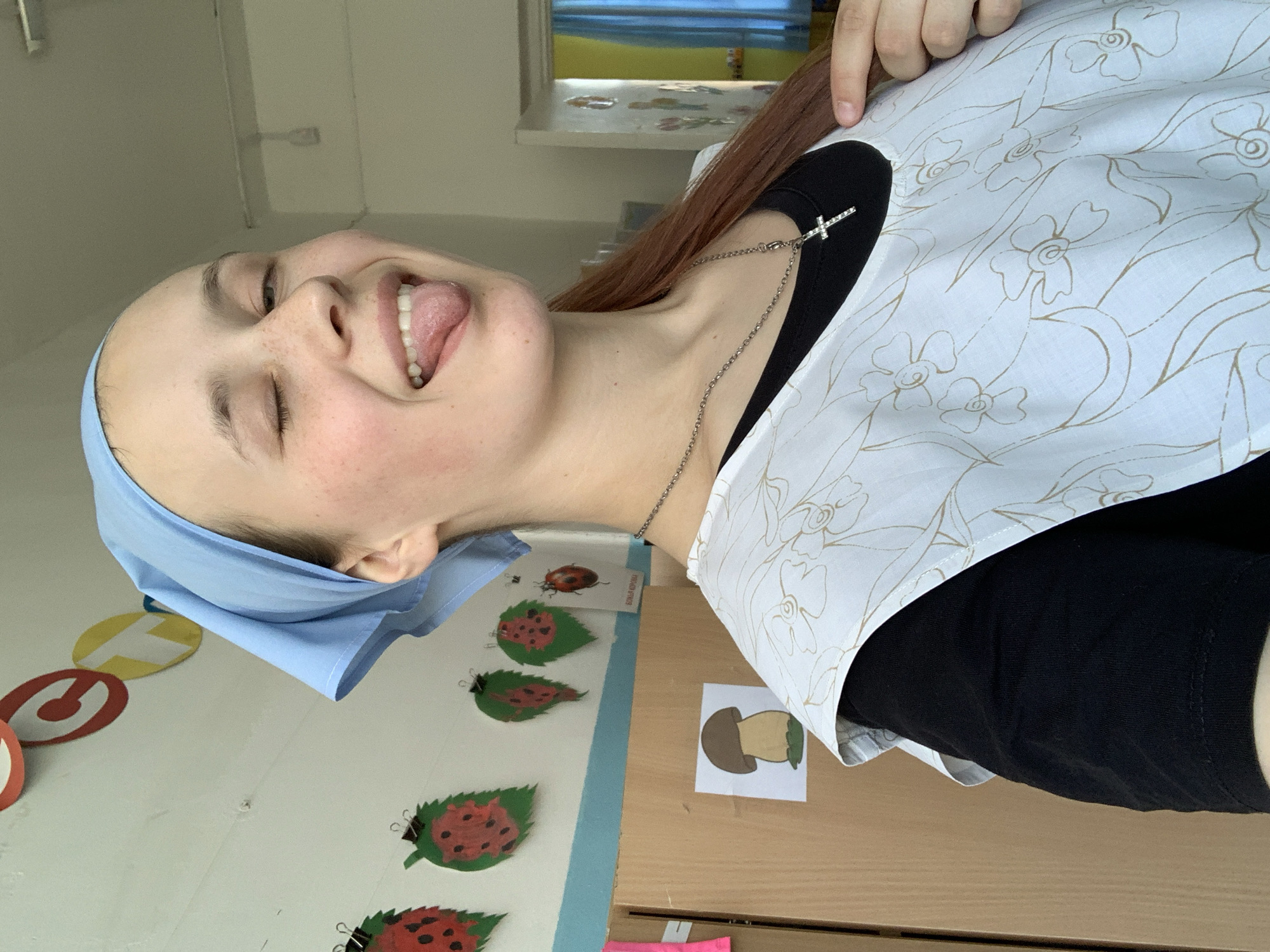 A woman with light skin, freckles, and a blue headscarf is sticking out her tongue and smiling with her eyes closed. She wears a black shirt under a white apron with a floral pattern, and a silver chain necklace with a cross. She is located in a room in Фоки, Russia, evidenced by text in Russian on a paper on the wall. The room has a wall with colorful circular decorations and paper ladybugs attached to leaves. A picture of a mushroom is attached to a desk.