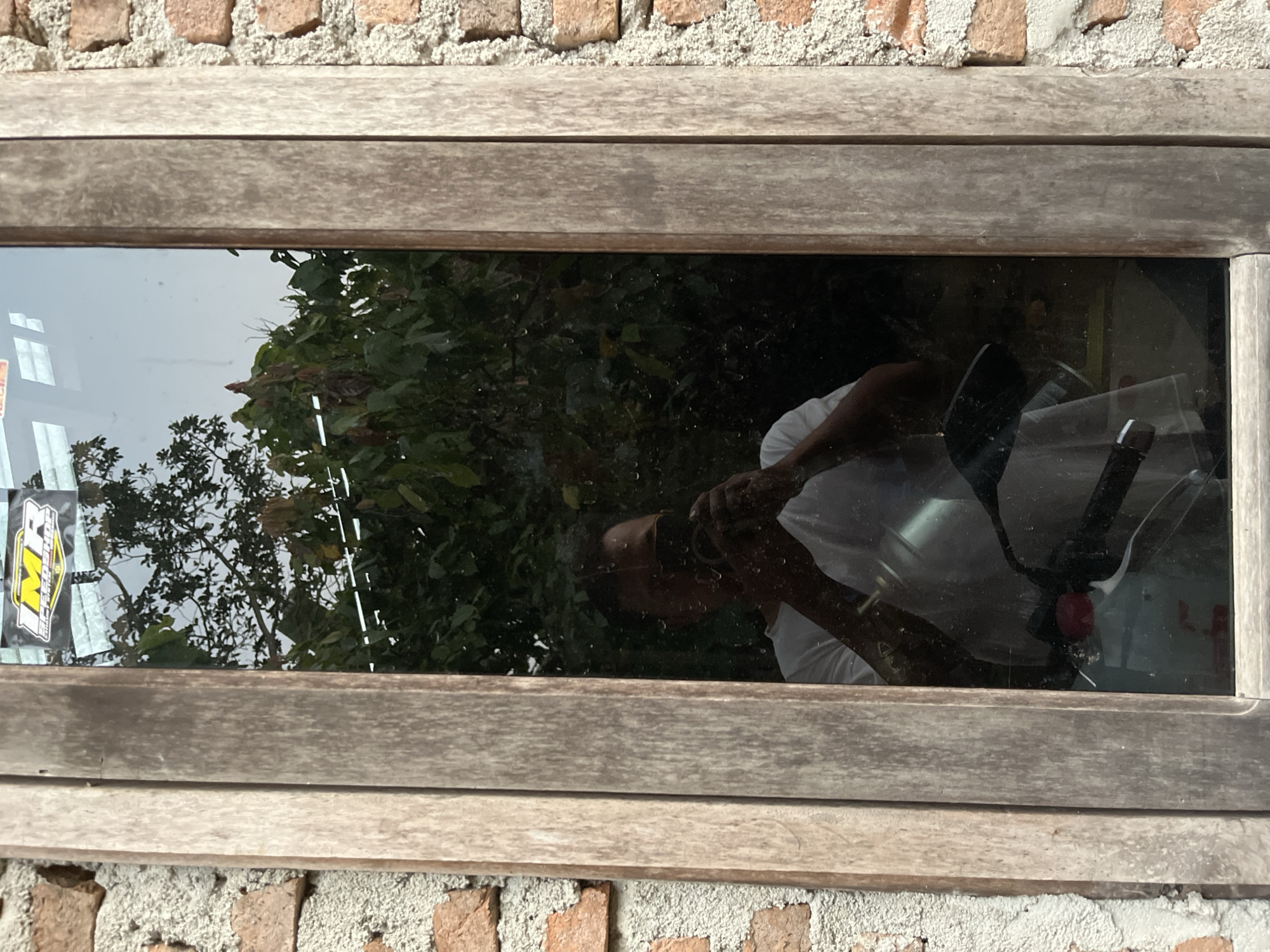 This is a vertical, eye-level, medium shot of a man taking a selfie in a window. The man is wearing a white t-shirt and is holding a black cell phone in his right hand, taking a picture of himself. He is reflected in the window, which is framed by weathered wooden planks. The window is set into a brick wall. Behind the man, green foliage and a bright sky are visible through the window. A sticker with the text "IMR RACING" is affixed to the top left of the window. The scene appears to be in a residential area in the city of Kebumen, Indonesia, given the brick construction and tropical foliage.