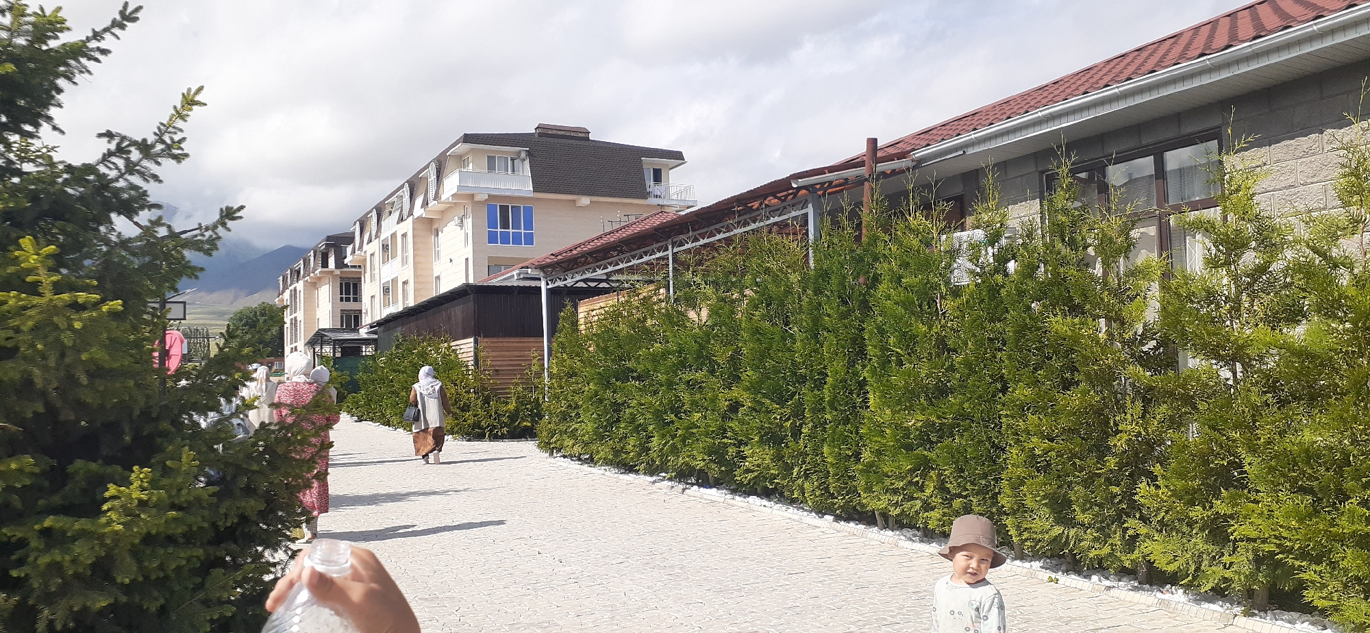 The image depicts a paved walkway in front of modern multi-story buildings, likely in the city of Choktal, Kyrgyzstan. A line of neatly trimmed evergreen trees borders the walkway, with a low white gravel border. In the foreground, a young child wearing a hat stands on the walkway, facing the camera with a neutral expression. To the left, a person's hand holding a clear plastic bottle is visible at the edge of the frame. Further down the walkway, two women are walking away from the camera. The woman on the right wears a light-colored headscarf and dress, carrying a dark bag. The woman on the left wears a patterned pink and white dress and a white headscarf. The background features a row of buildings with light-colored facades and dark pitched roofs. Mountains are visible in the distance under a cloudy sky.