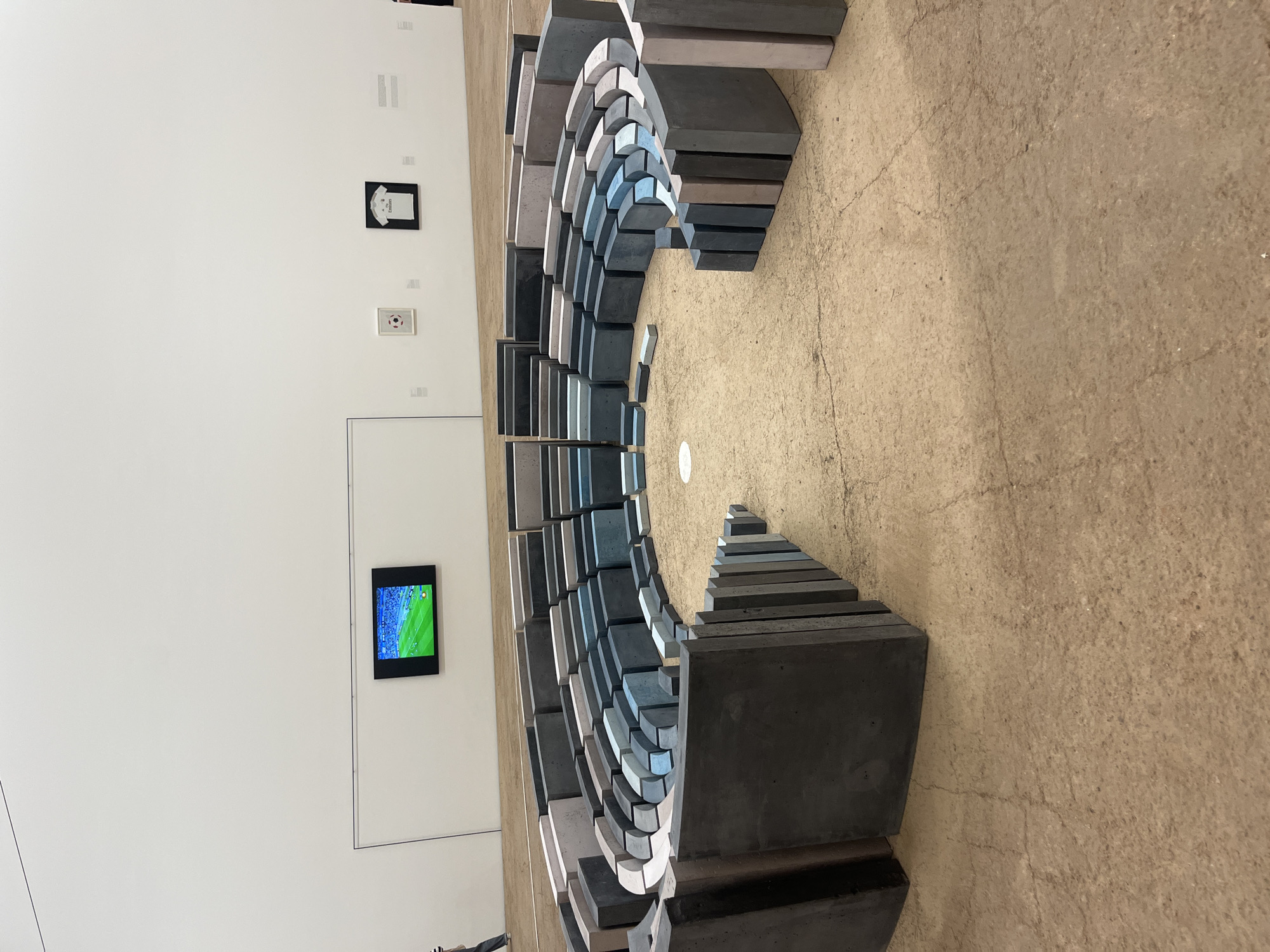 The image shows a large, circular installation of concrete blocks resembling an amphitheater in a museum or art gallery. The blocks vary in size and color, with shades of gray, blue, and beige. Some of the blocks are curved, creating a stadium-like seating arrangement. In the background, there is a white wall with a television screen displaying a green field, possibly a soccer game, and two framed artworks. The floor is covered in a light brown, textured material, possibly sand or concrete. The overall scene suggests an indoor exhibition space. The image does not provide any information about the location being Mexico City, Mexico.