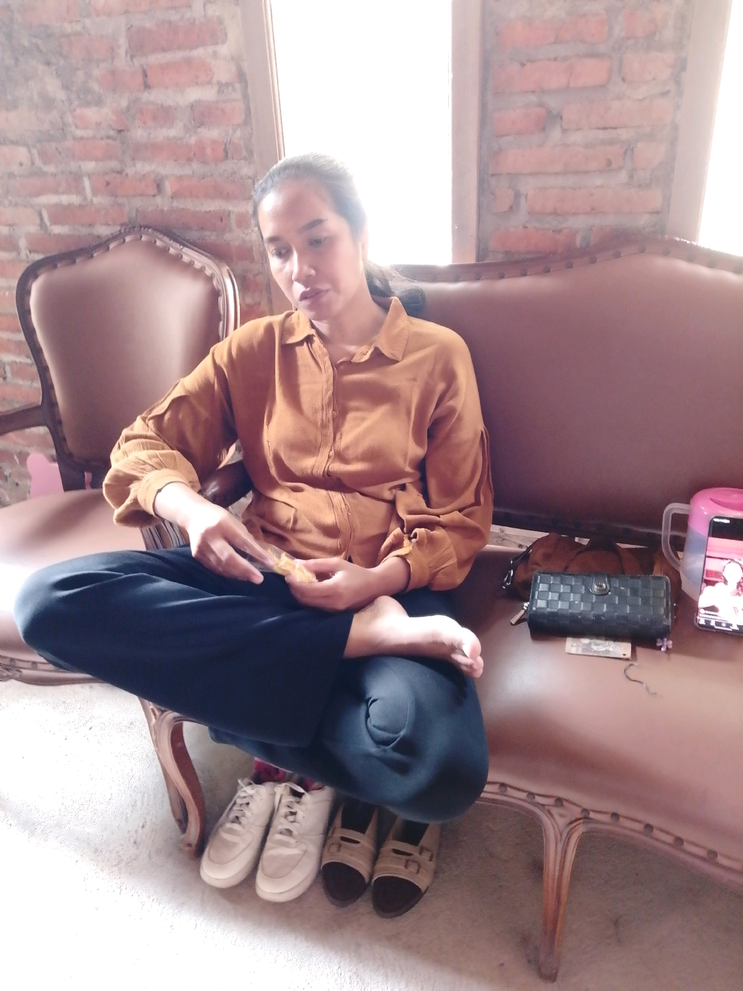 A woman sits on a brown sofa, facing slightly left, in what appears to be an indoor setting with a brick wall in the background. She is wearing a long-sleeved, collared, ochre-colored shirt and dark blue pants. Her legs are crossed, with her right leg resting on top of her left. She is holding a small, light-colored object in her hands, which are resting on her lap. Her gaze is directed downwards towards the object.
To her right, on the sofa, are several items: a dark brown purse, a black wallet with a checkered pattern, a small stack of bills or papers, a small purple flower-shaped object, a light pink pitcher, and a smartphone displaying an image of people.
On the floor in front of the sofa are four pairs of shoes: white sneakers, beige sandals, and another pair of brown sandals.
The lighting in the image suggests a bright environment, possibly with natural light coming from a window or opening to the right. The overall scene depicts a relaxed moment, possibly at home or a similar casual environment in Cikal, Indonesia.
