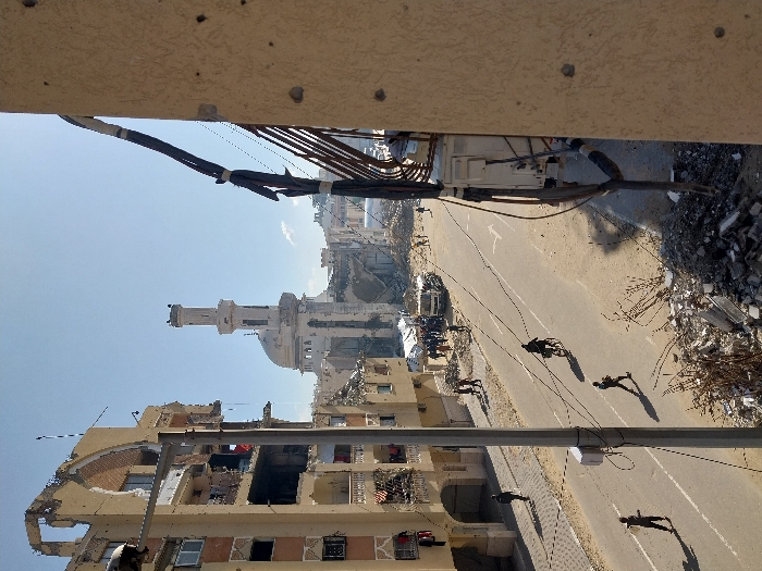 This image captures a wide shot of a severely damaged urban street in Khan Yunis, Palestinian Territory, under clear daylight conditions.
The scene is dominated by widespread destruction. To the left, a multi-story residential building exhibits significant structural damage, with large sections of its facade missing, exposing multiple interior rooms. Numerous impact marks or bullet holes are visible on its remaining exterior walls, and laundry is hanging from some balconies.
In the center-background, a prominent mosque with a minaret and a dome stands heavily damaged, showing partial collapse and extensive disfigurement to its structure.
The street, appearing to be paved, runs through the center, partially covered with sand and debris. White road markings, including a directional arrow, are visible on the asphalt. Approximately 15 individuals are dispersed across the street and its sides. One person is riding a bicycle in the middle of the road. Another individual walks carrying an object. A small group of people (around 10) are gathered near a white cargo truck and what appears to be a temporary tent structure further down the street.
In the immediate foreground on the right, a section of a building facade, likely the vantage point, displays multiple circular impact marks, consistent with bullet holes, and exposed electrical conduits with bundles of wires strapped to the wall. Piles of rubble, twisted metal, and assorted debris are scattered throughout the foreground and along the sides of the street. A utility pole with attached wires stands vertically in the left mid-ground.
