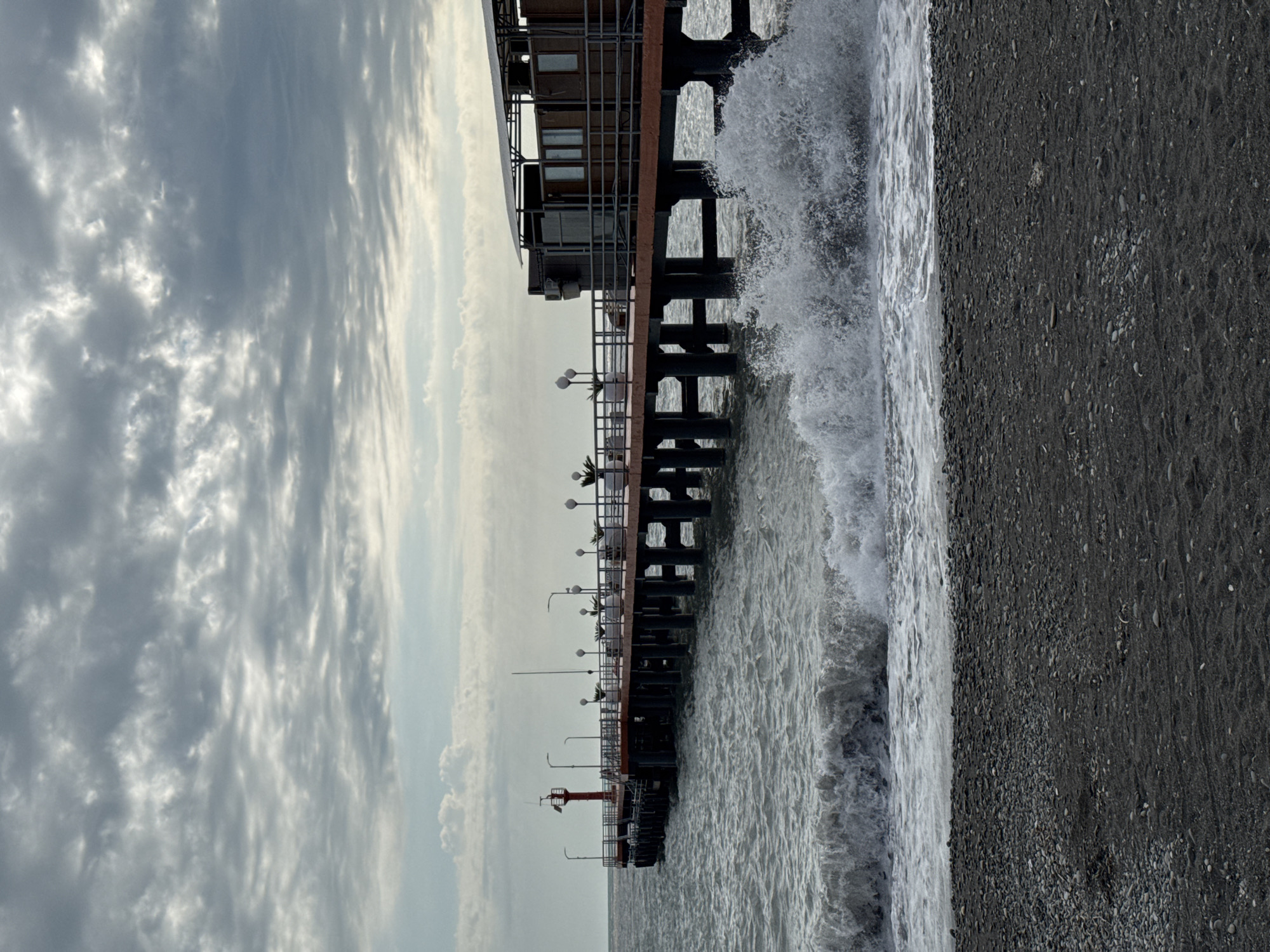 Here's a detailed description of the image:
The image captures a sideways view of a pier extending into the sea. The pier is constructed with dark-colored vertical supports and horizontal platforms, appearing sturdy and substantial. There's a building or structure situated at the beginning of the pier, possibly a restaurant or observation point. Smaller lamps line the side of the pier.
Waves are crashing forcefully against the supports of the pier, creating white foam and demonstrating the power of the sea. A dark, rocky beach occupies the right side of the image, where the waves meet the shore.
The sky is filled with gray and white clouds, suggesting an overcast day. The overall lighting is subdued.
Given the location information, this scene is likely depicting a pier along the Black Sea coast in Sochi, Russia.