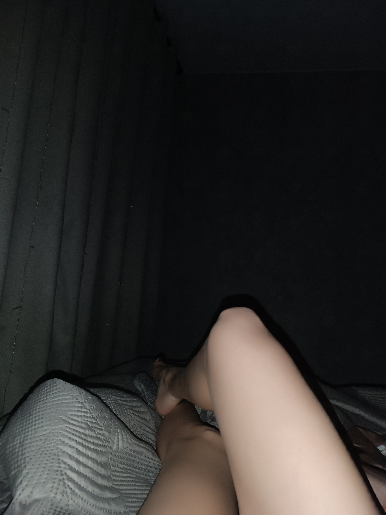 A close-up shot of legs resting on a bed with a grey quilted blanket. The location appears to be indoors, possibly a bedroom, with dark curtains visible in the background. The city is unknown.