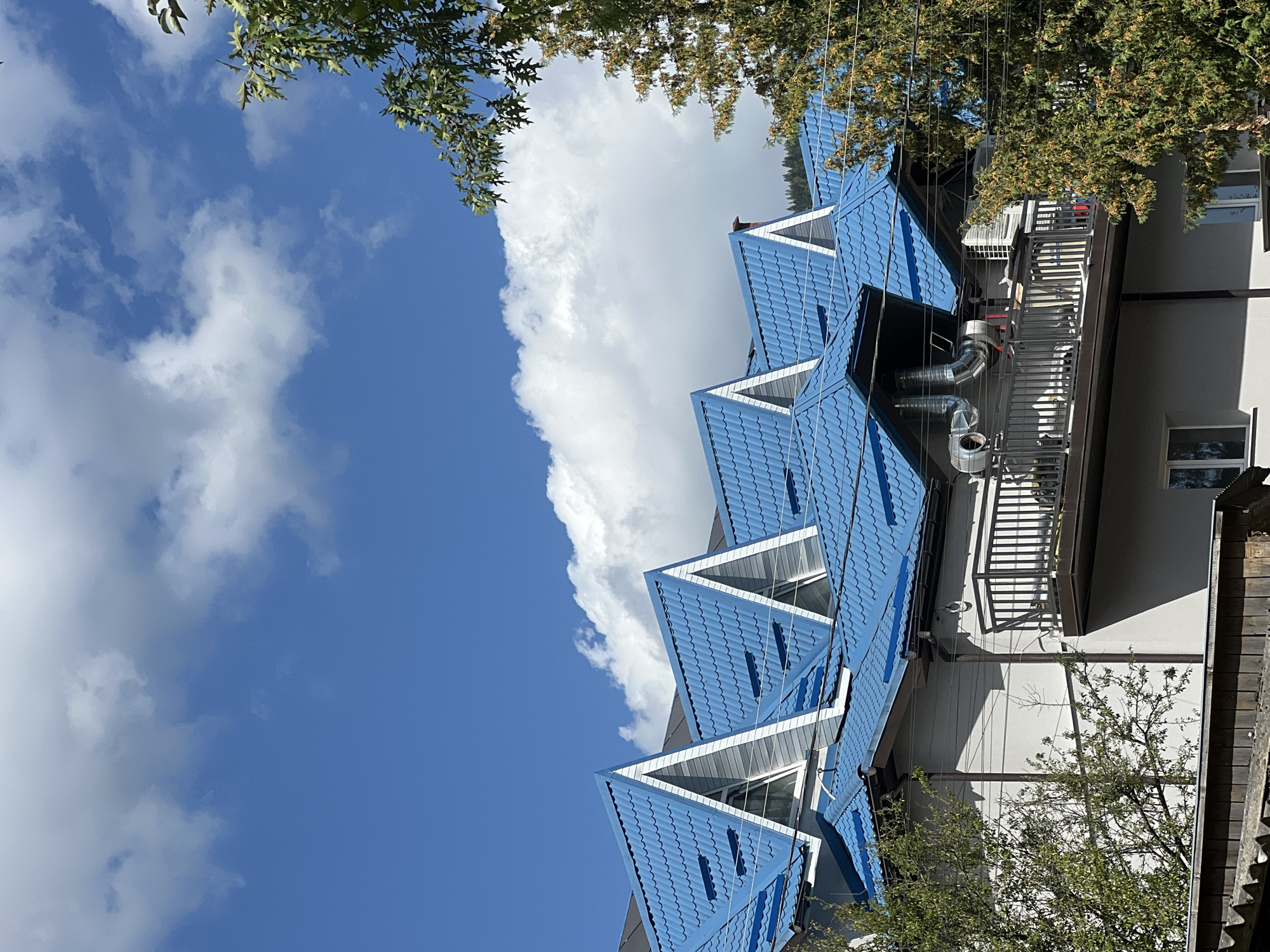 In Yaremche, Ukraine, this photograph captures a multi-story building with a striking blue, wave-patterned metal roof featuring prominent triangular gables. The building has a light-colored facade, a dark railing surrounding a balcony, and several windows. Two air conditioning units are visible on the balcony, along with large metal ventilation pipes. Trees with green foliage frame the building on the right and left sides, and a section of a wooden fence is visible at the bottom. The sky above is a vibrant blue, punctuated by fluffy white clouds. The image appears to be taken on a bright, sunny day.