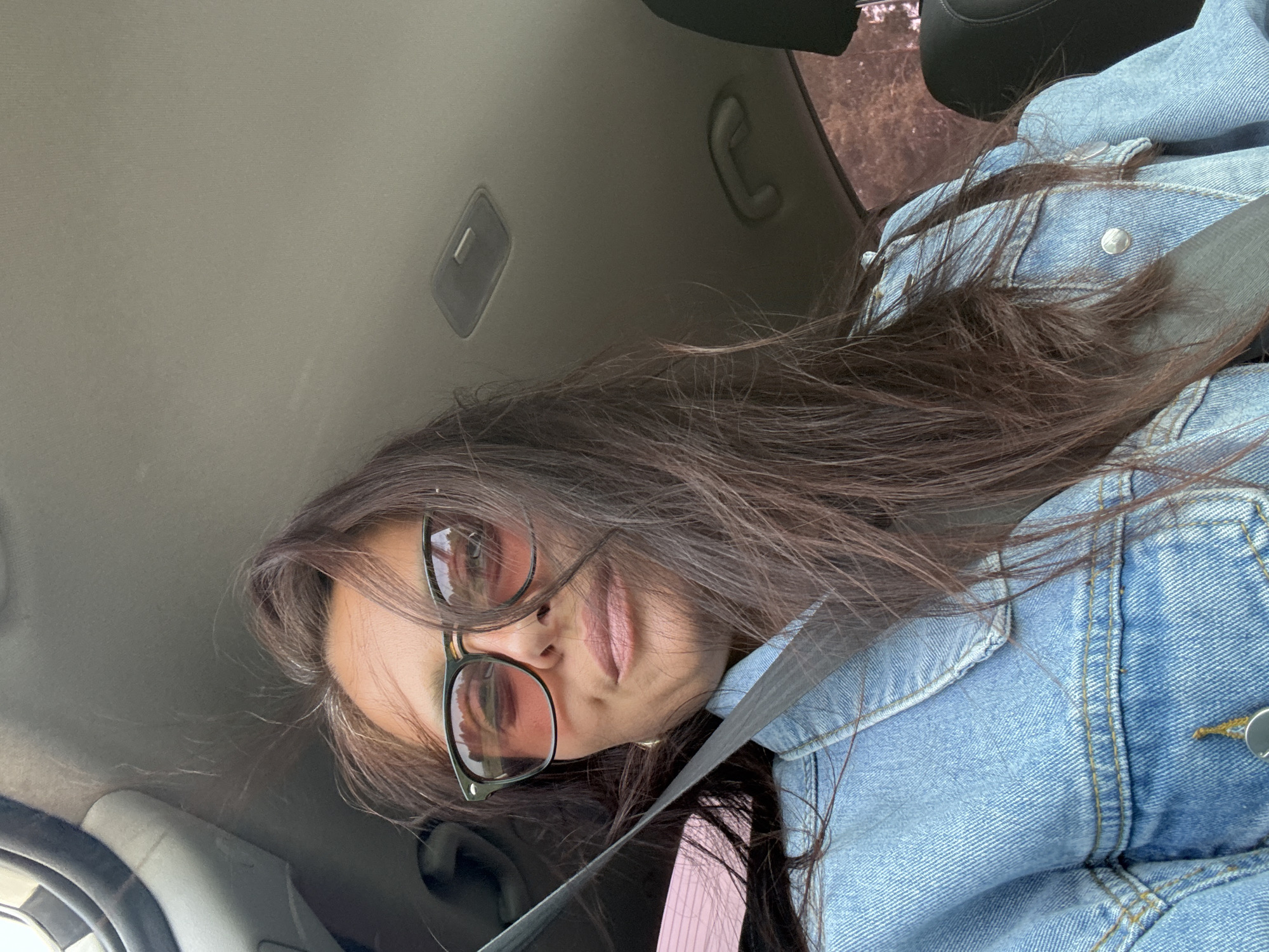 A woman with long brown hair and glasses sits in the driver's seat of a car, wearing a blue denim jacket and a seatbelt. The interior of the car is visible, including the roof lining and a portion of the steering wheel. The location is Chaikovsky, Russia.