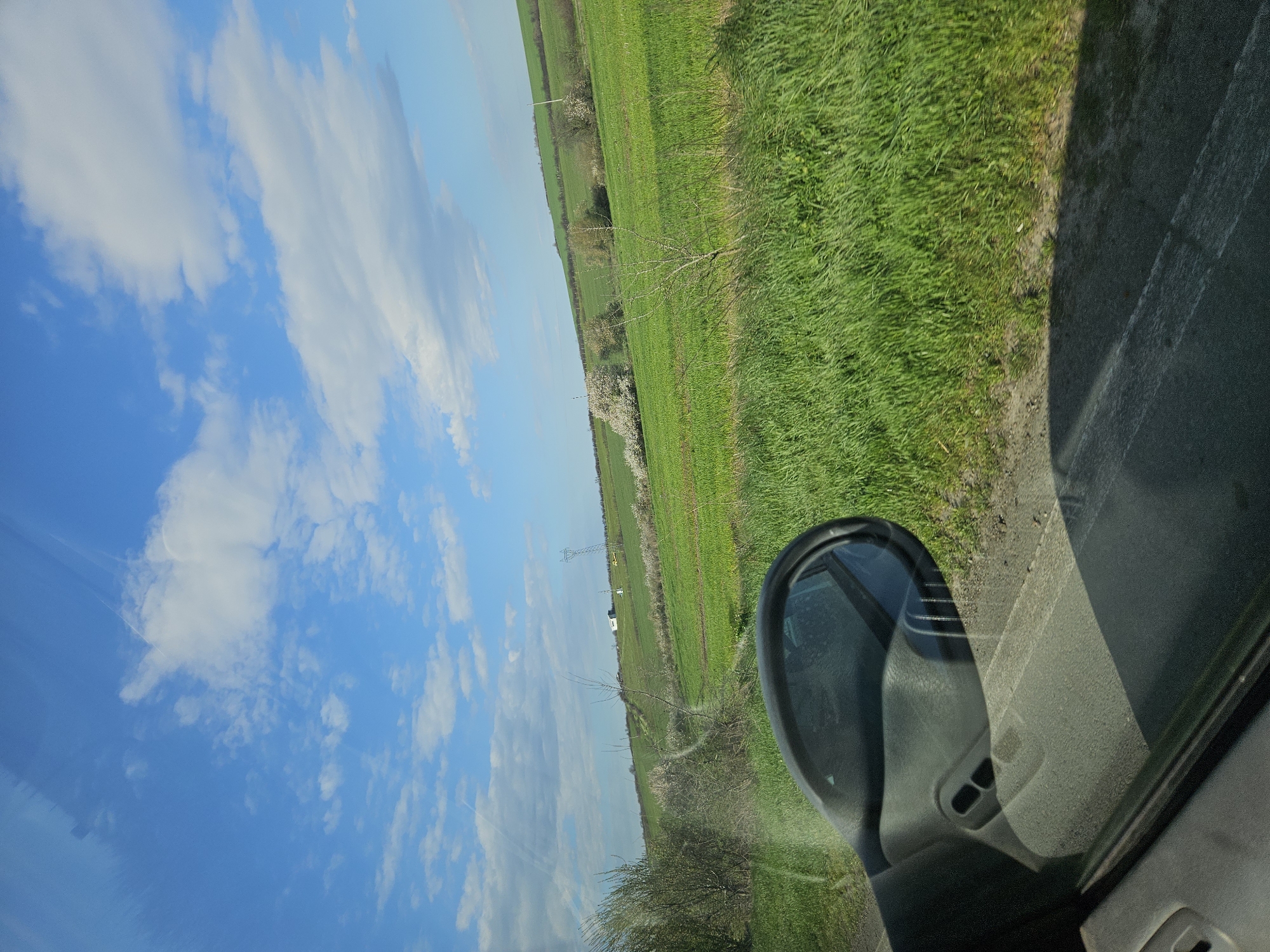The image depicts a view from inside a vehicle, likely a car, showing the side mirror, part of the interior, and the scenery outside. Outside, there is a field of green grass next to a road. Beyond the field, there appears to be a line of trees and possibly other vegetation. The sky is blue with scattered clouds. There is no indication this scene is within the city of Marinka, Bulgaria, or any specific location for that matter. There are no visible people or discernible actions occurring.