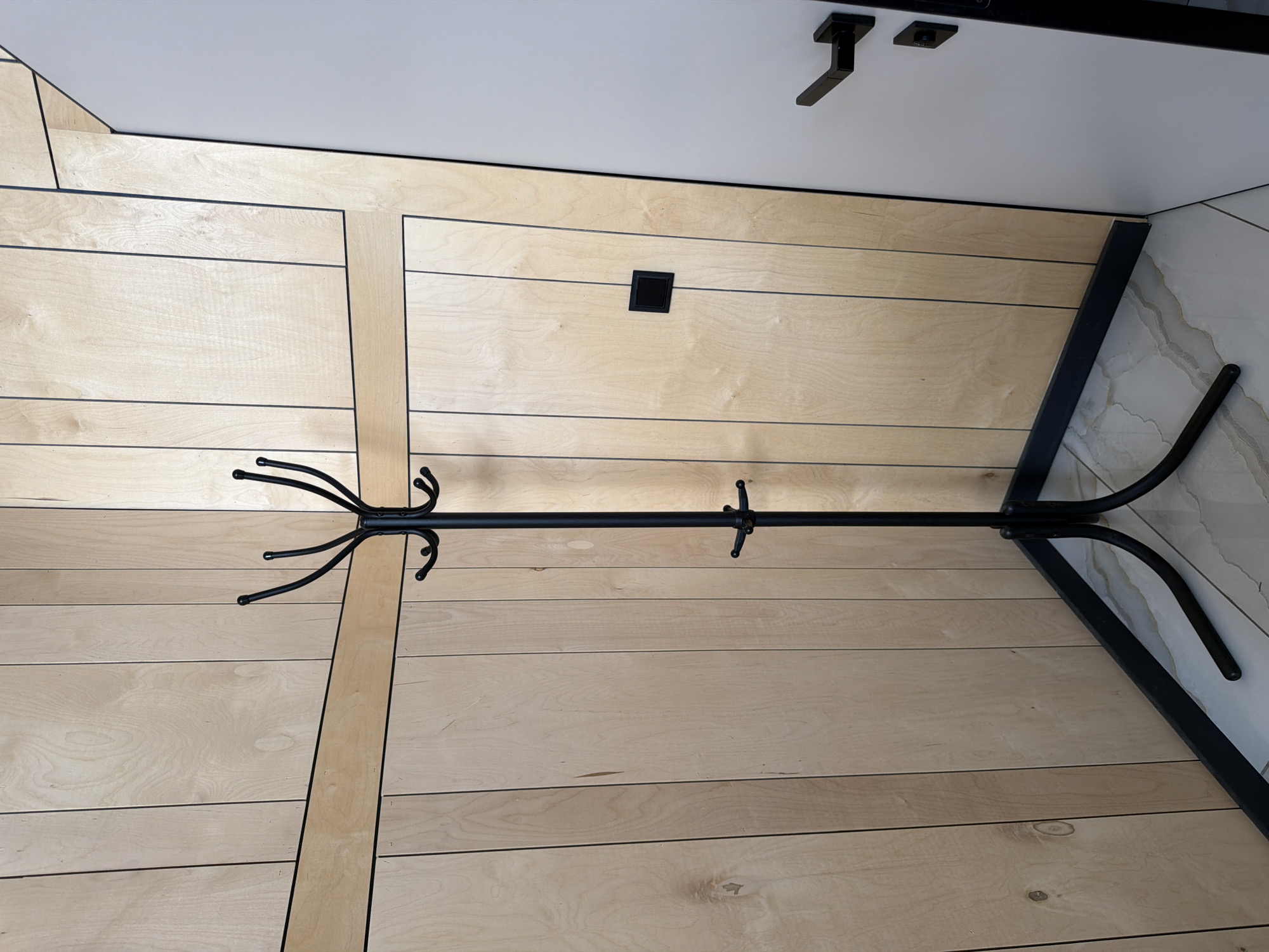 The image shows a black metal coat rack standing in a corner. The coat rack has a central pole with several hooks at the top and a smaller set of hooks lower down. The base of the coat rack is a tripod design with curved legs. The background is a wall paneled with light-colored wood, with darker horizontal and vertical trim lines. To the right of the coat rack, a portion of a white door with black hardware is visible. The floor appears to be tiled with a light-colored pattern. There is a small black square object mounted on the wood paneling to the right of the coat rack. The scene is lit from above and to the left. This image was taken in Семилуки, Russia.
