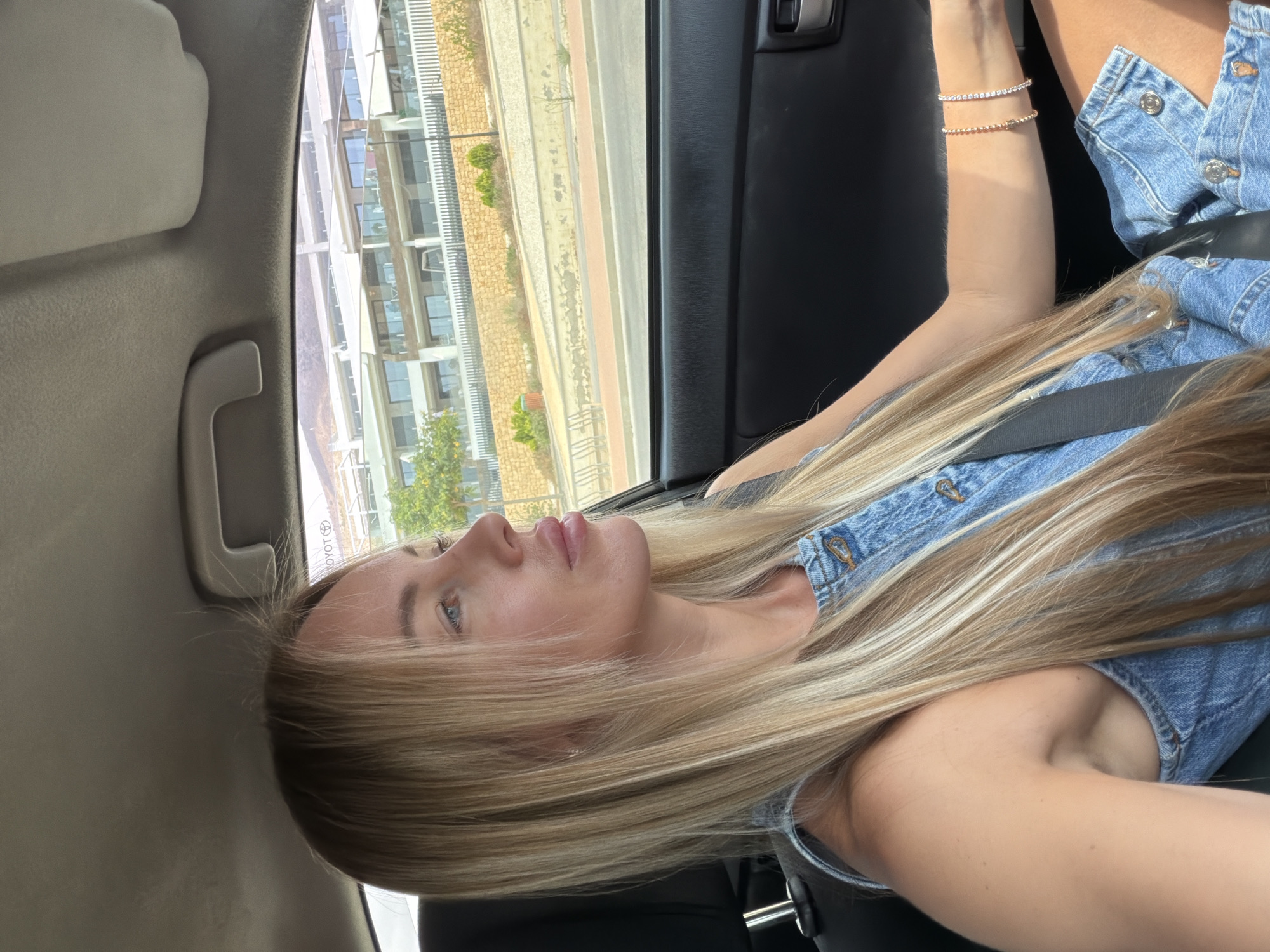 A blonde woman is sitting in the driver's seat of a car, looking out the window. She is wearing a denim dress and has blonde hair. Outside the car, there is a building with multiple balconies and a street with a sidewalk. The location is Finestrat, Spain. The car's interior is visible, including the dashboard and the driver's side door.
