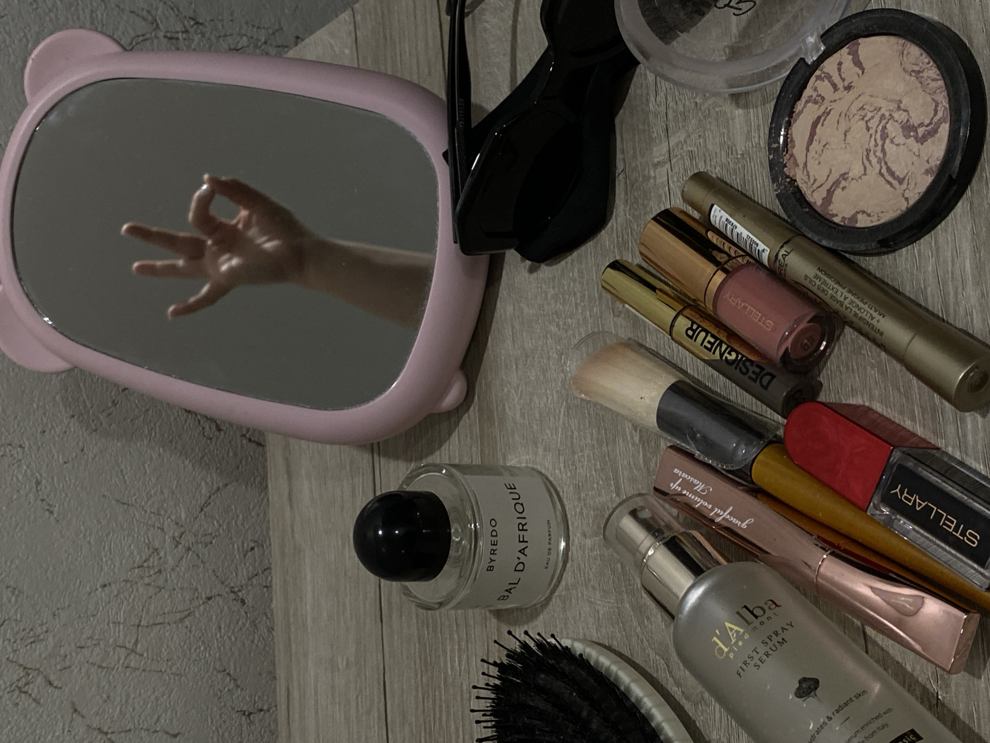 The image captures a flat lay of various personal care and cosmetic products arranged on a light wooden surface, possibly a vanity or table. In the upper left quadrant, a pink, oval mirror with what appears to be ear-like protrusions reflects a fair-skinned hand making an "OK" gesture. Behind the mirror, a section of a textured, greyish wall is visible.
Among the items scattered on the surface are:
- A compact powder with a marbled beige and brown pattern in a dark circular case.
- A golden tube of L'Oréal mascara, with visible text including "INTENSIFIE LA BASE DES CILS", "ALLONGE À L'EXTRÊME", and "MULTI-PEIGNE PRÉCISION".
- A pink lip gloss or liquid lipstick labeled "STELLARY".
- A tube marked "DESIGNEUR" and another pink metallic mascara tube with "graceful volume up Mascara" text.
- A black rectangular bottle of "STELLARY" branded product, possibly eyeliner.
- A clear glass bottle of "BYREDO BAL D'AFRIQUE EAU DE PARFUM" perfume with a black cap.
- A silver pump bottle of "d'Alba piedmont FIRST SPRAY SERUM", which highlights "hydrates & radiant skin" and mentions being "from Italy".
- Two makeup brushes (one large, one smaller with a red handle) and a pair of folded black sunglasses.
- Part of a black hairbrush is visible at the bottom left.
The arrangement suggests a collection of daily beauty essentials, possibly set out for use or after use. The ambient lighting indicates an indoor setting. The reflected "OK" gesture adds a personal and positive touch to the scene, which is located in Sochi, Russia.