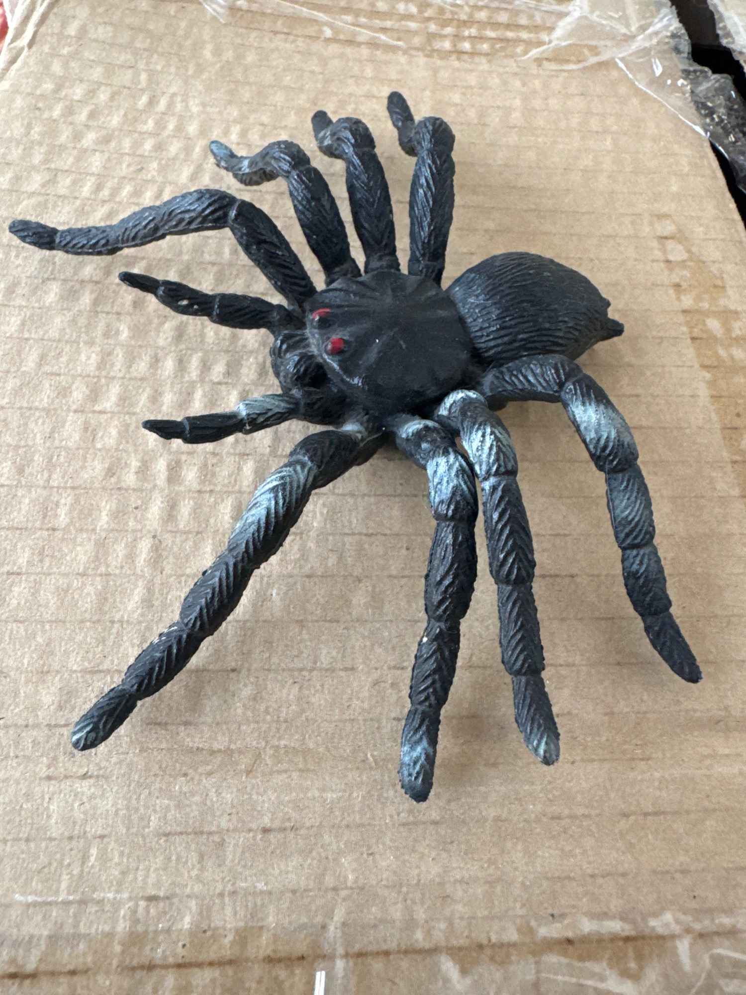 A black plastic spider toy with red eyes is positioned on a brown cardboard surface. The spider has eight segmented legs, with some of them appearing lighter colored due to wear or detailing. The toy is lying on its back. The background is a textured cardboard surface, with some faint lines or corrugations visible. There is a piece of clear plastic sheeting visible in the upper right portion of the image. The location is not determinable from the image.