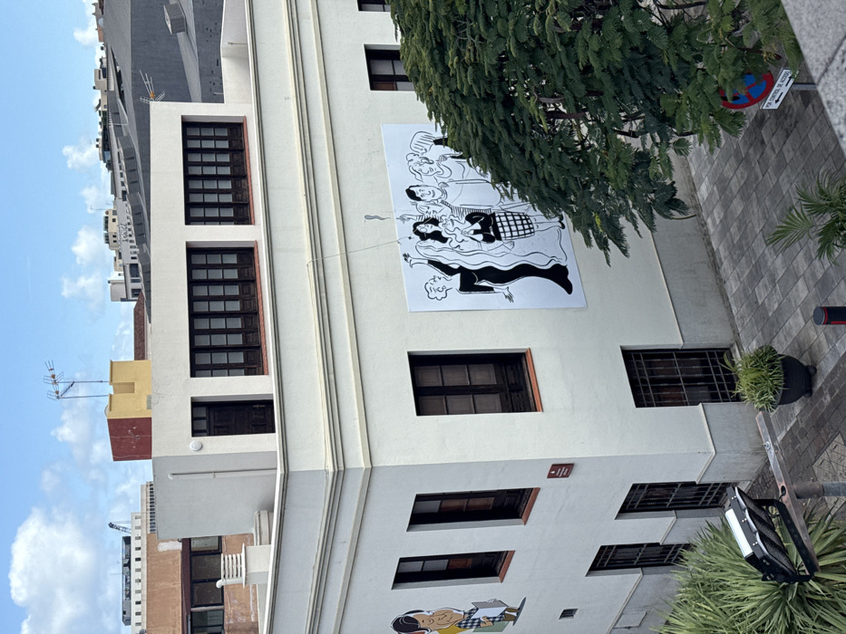 A tall, light-colored building features a large graphic mural of cartoon-like figures on its upper facade, adjacent to a tree. Dark-framed windows are visible on multiple levels. The surrounding area includes tiled pavement, a street light, greenery in pots, and a no parking sign. Other buildings and a blue sky with clouds are in the background.