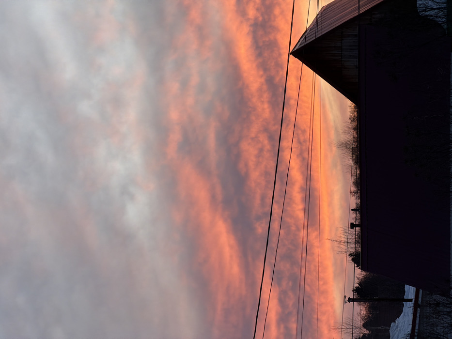 The image depicts an outdoor scene at either dawn or dusk in the city of Chaikovsky, Russia. The sky dominates the upper two-thirds of the frame, displaying a transition from light grey and white at the top to vibrant orange, pink, and red hues towards the horizon, indicative of a sunrise or sunset. Clouds are scattered across the sky, intensely illuminated by these colors.
Multiple dark, thin utility lines run horizontally across the mid-section of the image. Below these lines, in the lower third of the frame, structures are silhouetted against the bright sky. On the left, a dark utility pole is visible, adjacent to the dark-colored roof of a building, which appears to be dark red or maroon and features two small, dark, chimney-like protrusions. To the far left, a patch of white ground cover, consistent with snow, is present. Bare tree branches are silhouetted above the roofline. On the right side, the prominent dark silhouette of an A-frame building with a steeply pitched roof, possibly constructed of wood, rises into the frame. No individuals are visible, and no specific actions or interactions are discernible. The environment suggests a suburban or semi-rural setting within the specified city.