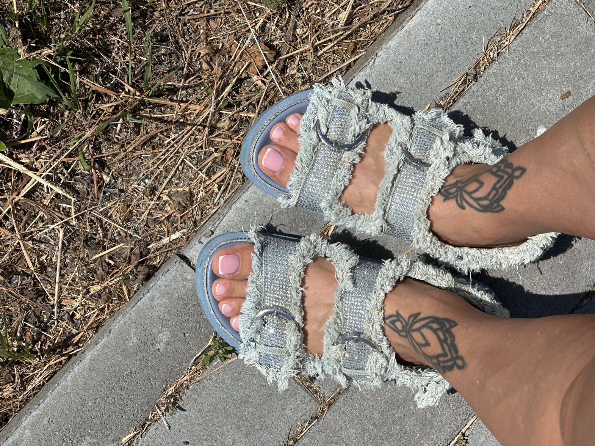 A pair of feet are shown from a top-down perspective, resting on a concrete pathway adjacent to dry grass and some greenery. The feet are adorned with light blue, distressed denim sandals, each featuring two buckled straps embellished with rhinestones and a frayed edge. The toenails are painted a pale pink with white tips, giving them a French manicure appearance. Each foot has a black tattoo on the ankle area; the left foot displays a tattoo resembling a stylized geometric knot or emblem, while the right foot has a similar but distinct geometric design. The scene is lit by bright sunlight, casting subtle shadows. The environment appears to be an outdoor setting, possibly a park or a sidewalk in the city of Yuzhnouralsk, Russia, given the contrast between the paved walkway and the natural, somewhat arid vegetation.