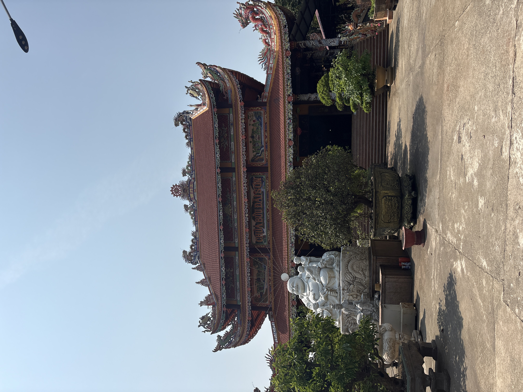 The image displays an ornate Buddhist temple or pagoda in Đà Nẵng, Vietnam, under a clear, bright blue sky. The main subject is a multi-tiered building featuring a traditional red tiled roof with steeply angled eaves. The rooflines and facade are extensively decorated with intricate carvings, including numerous dragon figures, auspicious symbols like the Dharma wheel, and detailed patterns in vibrant colors. Murals or paintings are visible on the temple's walls between its levels.
In the foreground, to the right of the temple, stands a large, white stone statue of the Laughing Buddha (Hotei/Budai), portrayed with several small child-like figures climbing on him. This statue is placed on a finely carved base, which is part of a larger, decorative white stone structure. Various green trees and potted plants are positioned around the statues and the temple entrance, contributing to the tranquil atmosphere.
The ground is a paved concrete or stone courtyard that shows some cracks and weathering. Strong sunlight bathes the scene, casting distinct shadows from the foliage and architectural elements, suggesting it is midday or early afternoon. A modern street light fixture is partially visible against the clear sky in the upper left corner.
The visible text on the side of the temple building reads "CHÙA BỬU ĐÀI SƠN," which translates to Buu Dai Son Pagoda. The scene is static, devoid of people or explicit activity, focusing on the architectural beauty and spiritual essence of the temple grounds.