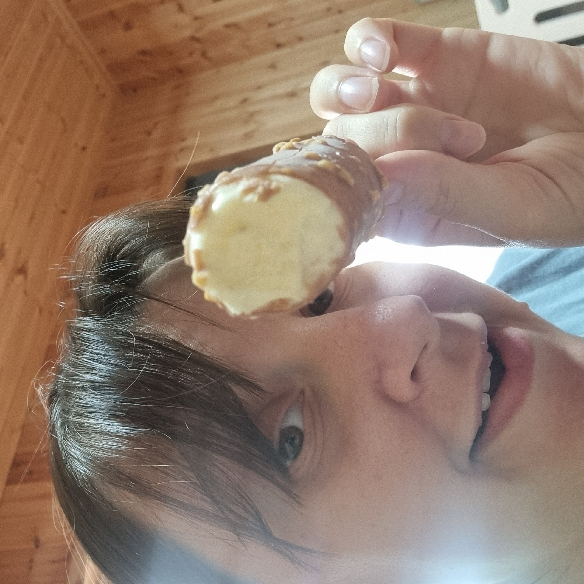 In this close-up, high-angle shot, a person is holding an ice cream cone up to the camera, obscuring part of their face. The ice cream is partially eaten, revealing a white, creamy interior and a chocolate coating with visible nut pieces. The person has brown hair with some lighter strands, and their skin appears to be fair. Their mouth is open in a smile, showing teeth. The background consists of a wooden wall, suggesting an indoor setting. The image captures a moment of enjoyment, with the focus on the treat. The specific location is not identifiable in the provided image.