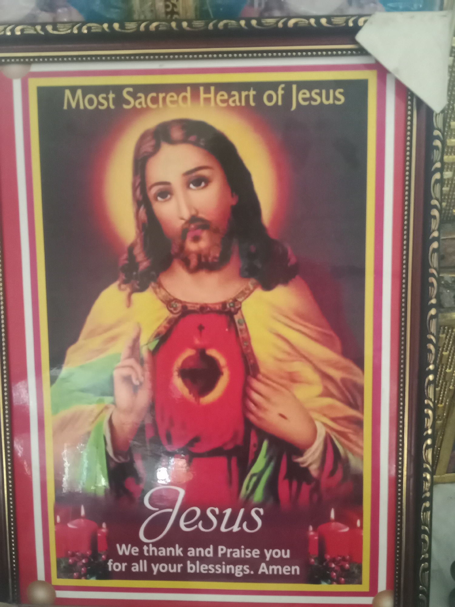 A framed portrait of Jesus Christ with the inscription "Most Sacred Heart of Jesus." Beneath the image of Jesus, in cursive font, is the name "Jesus" followed by the phrase "We thank and Praise you for all your blessings. Amen." The image is bordered with a red, white, and yellow border, and framed in a dark brown and gold ornate frame. The location is Lokoja, Nigeria is not discernible from the image.