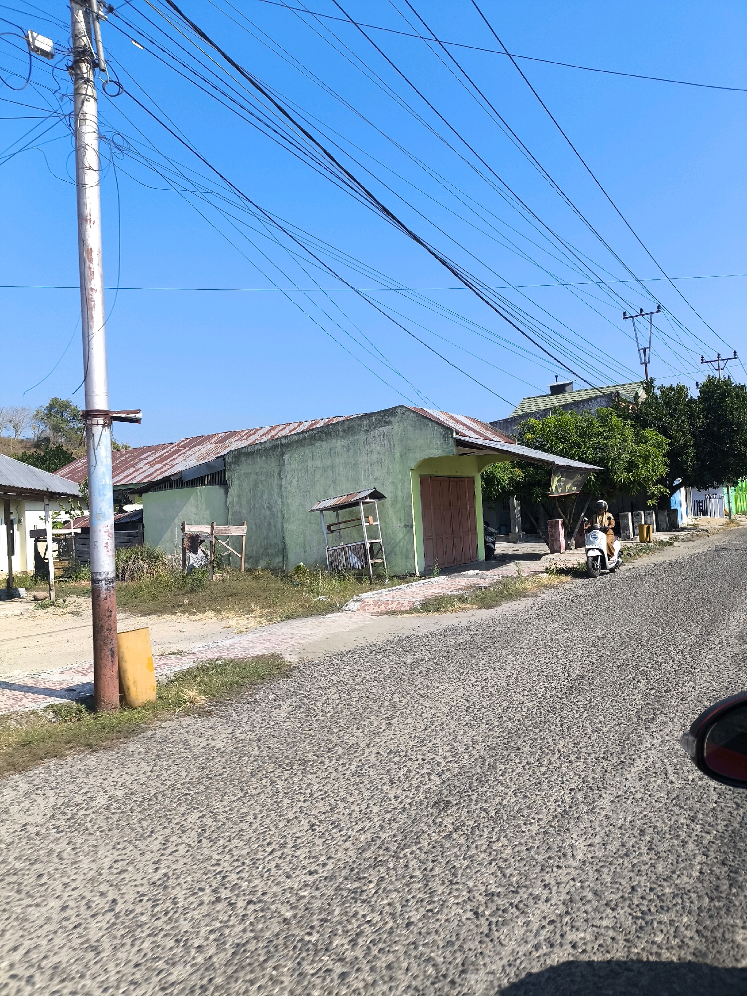 This image captures a street scene in Tilamuta, Indonesia, under a clear blue sky. A power pole, adorned with numerous black electrical wires, stands prominently on the left side of the frame. Adjacent to it is a yellow cylindrical object, likely a warning marker or a post.
The foreground is dominated by a gray asphalt road with a textured, pebble-like surface. To the right of the road, a portion of a red car's side mirror is visible, indicating the photographer's perspective.
The middle ground features a row of modest buildings. The most prominent is a light green building with a rusty, corrugated metal roof and a large, light brown double door. In front of this building, a white scooter is parked, and a person wearing a light brown outfit is seated on it. To the left of the green building, a smaller white building with a metal roof is partially visible. In between these two structures, the ground is covered with dry grass and some debris, including a wooden structure that appears to be a frame or a makeshift shelter.
In the background, more buildings and lush green trees line the street. The presence of utility poles with electrical lines suggests a developed, albeit possibly rural or semi-urban, area. The overall impression is one of a sunny day in a local Indonesian town, with everyday life unfolding on the street.