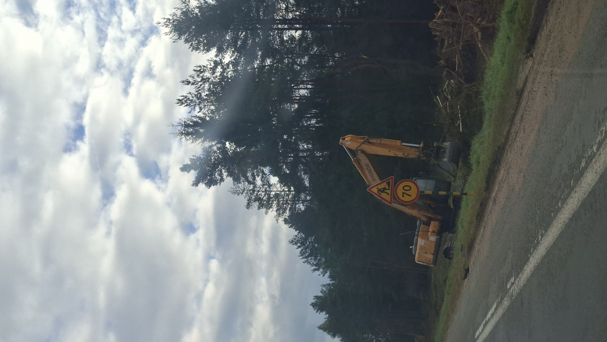 The image captures an outdoor scene featuring a yellow Hyundai excavator parked on a grassy verge next to a paved road. The excavator is positioned near a dense forest of tall pine trees. A traffic sign displaying "70" inside a circle is visible in front of the excavator, along with a triangular warning sign depicting a person with a shovel. The sky is partly cloudy, with a mix of gray and white clouds. The ground around the excavator is disturbed, with piles of branches and debris suggesting ongoing work. The road appears to be wet, with a double white line marking the lane. The overall environment suggests a construction or forestry site in a rural area, potentially along a roadway bordering a forest. The location is identified as Бягомльскі сельскі Савет, Belarus.