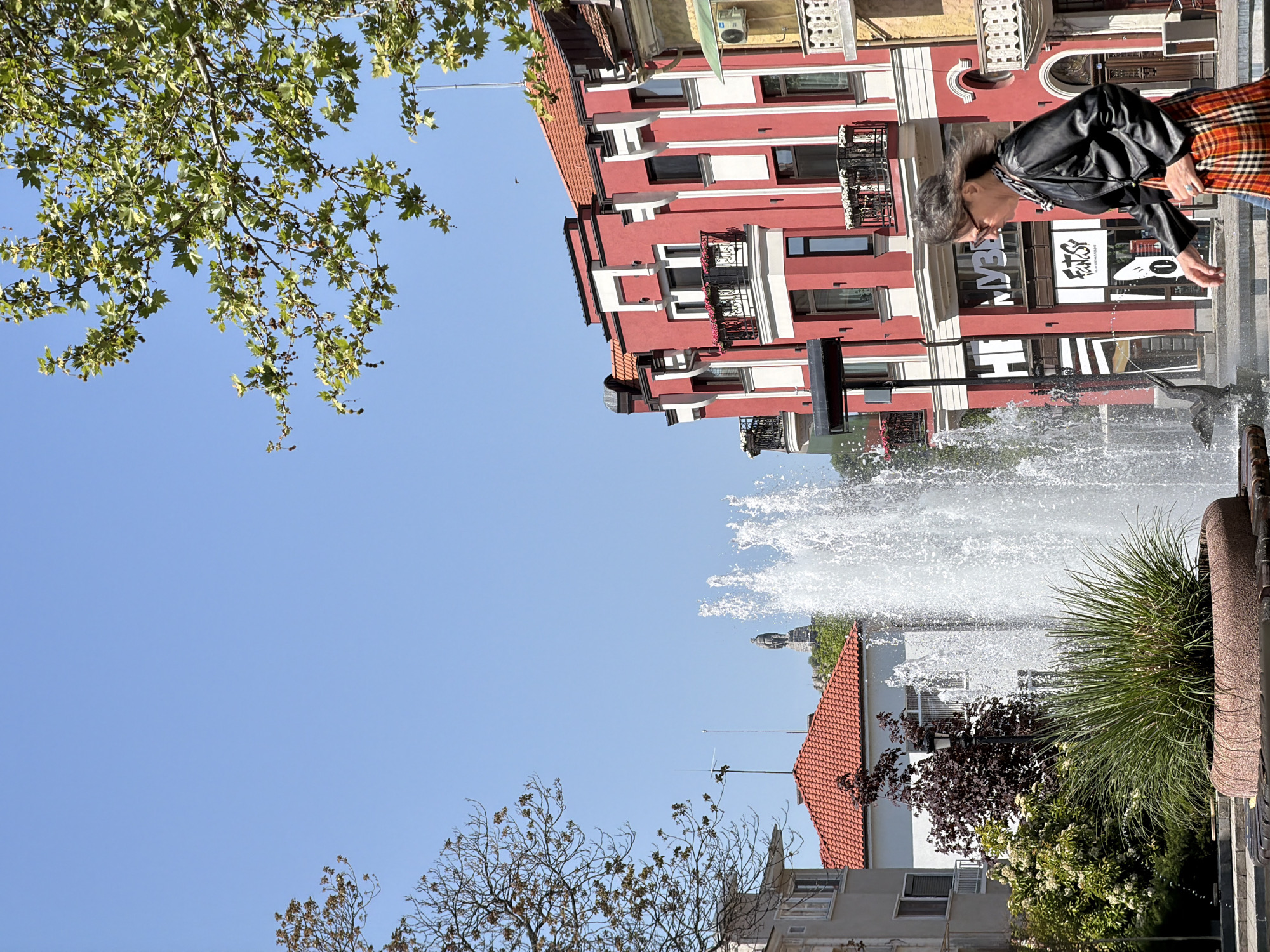 This image presents a sunlit urban scene in Plovdiv, Bulgaria, under a clear blue sky. The composition is vertically oriented, with significant sky space on the left and upper left, and architectural and natural elements filling the right and bottom right.
A prominent red multi-story building with white trim, multiple windows, and decorative balconies, stands on the right side of the frame. Its ground level features large display windows with visible text: "FANTASY" and "24", along with partial Bulgarian text including the letters "M" and "УВ". In front of this building, an active fountain sprays jets of water upwards, forming a bright arc.
To the right of the fountain, an adult person is visible from the side, wearing a black leather jacket and what appears to be an orange and black plaid skirt or trousers. They have dark hair and are looking downwards towards the fountain or an area to its left.
The upper left corner is filled with the green, lobed leaves and seed balls of a plane tree, illuminated by sunlight. The lower left and background reveal another building with a reddish-tiled roof and white walls, surrounded by various trees and bushes, some bare and others evergreen.
The scene depicts a bright, sunny day, likely late morning or afternoon, with strong natural light creating visible shadows. The overall atmosphere is calm, capturing a moment of everyday life in a public square or street.