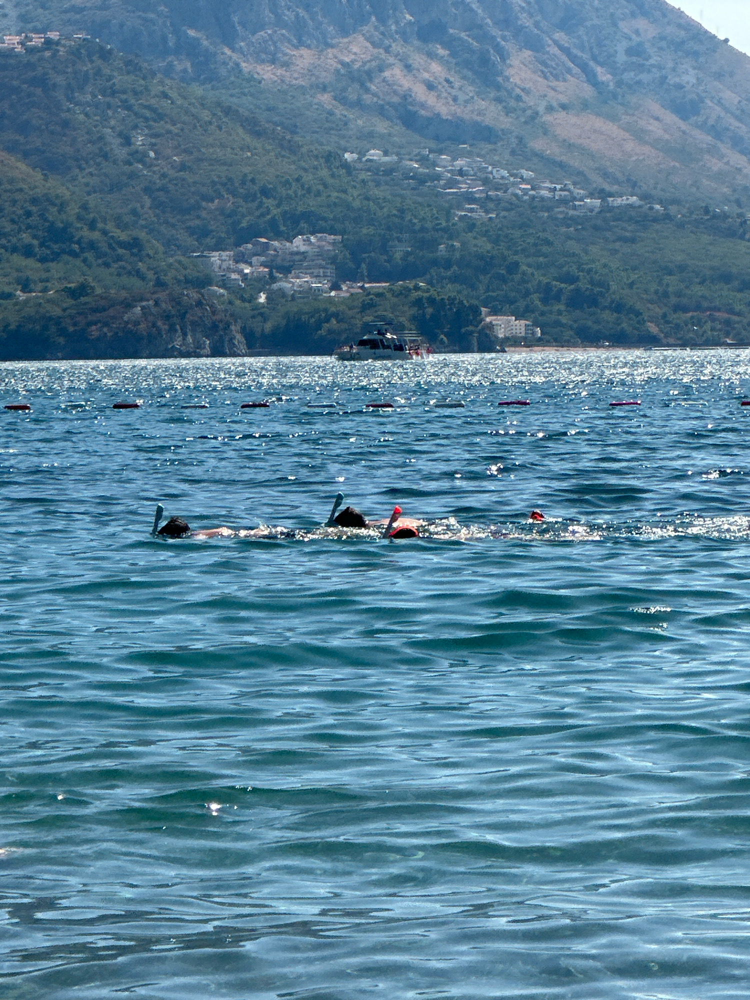 The image depicts a body of water with several people snorkeling. In the foreground, two individuals are visible, each with a snorkel and mask, appearing to be swimming. Further out in the water, there are other snorkelers and buoys. In the background, a mountainous landscape with scattered buildings is visible, likely representing the city of Boreti, Montenegro. A boat is also present in the distance, on the water. The sea is sparkling under a sunny sky.