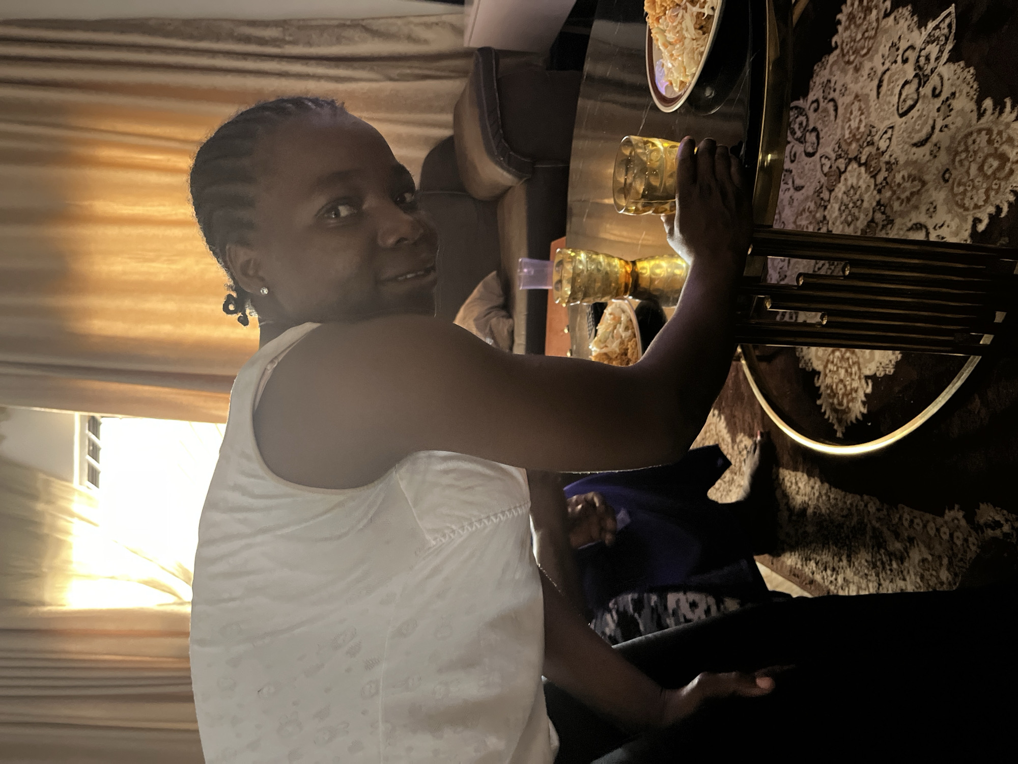 A dark-skinned woman with braided hair and a white tank top is looking over her shoulder at the viewer. She is seated at a dark, possibly marble table, set with food and drinks in golden, ornate glasses. A beige, patterned rug is visible beneath the table. The setting appears to be indoors, near a window with drawn curtains. The image does not provide sufficient information to determine the exact location in Jos, Nigeria.