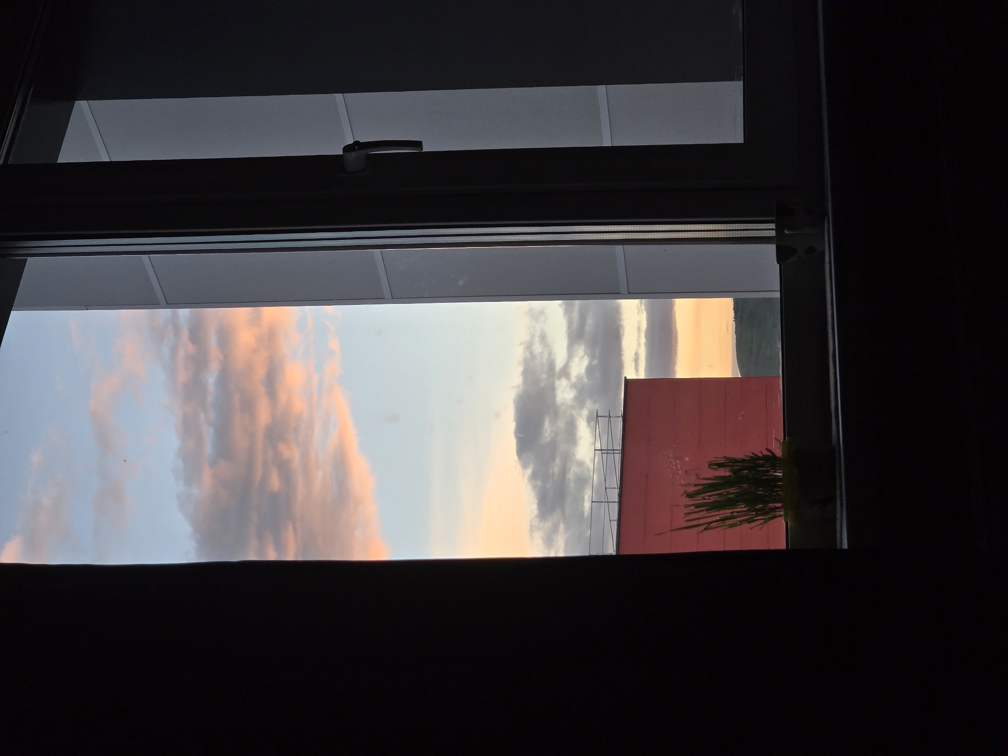 The image shows a view through a window at dusk. The sky is a gradient of blue, pink, and orange, with scattered clouds. In the foreground, a dark curtain or wall partially obstructs the view on the left side of the frame. The window itself has dark framing, and a handle is visible on the right side.
Outside the window, across a small gap between buildings, a flat-roofed building with a pink facade and a metal railing is visible. A potted plant with green, grass-like foliage sits on the windowsill in front of the pink building. Further in the distance, there are hints of a hilly landscape and other buildings. The overall scene suggests an urban or suburban environment at the end of the day.