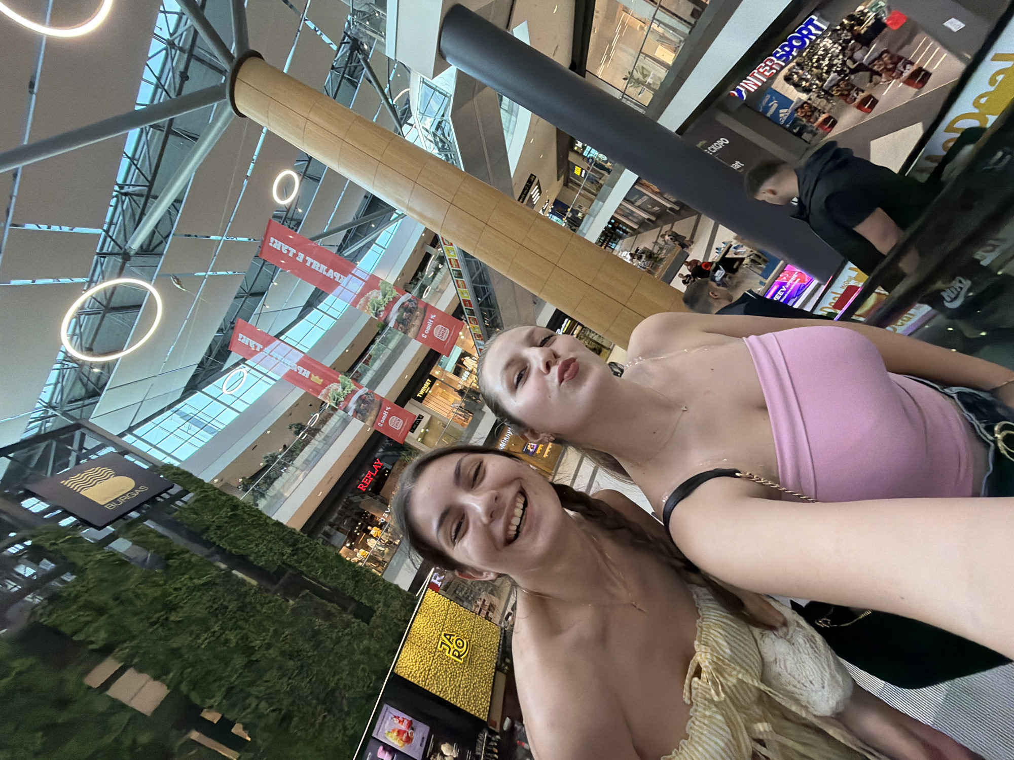 The media file presents a dynamic, wide-angle shot of two young women posing for a photo inside a modern, multi-level shopping mall. The image is taken from a slightly low angle, looking upwards and across the spacious interior. On the right, a woman in a pink strapless top looks towards the camera with a "duck face" expression. To her left, another woman with braided hair smiles broadly.
The background reveals the expansive architecture of the mall, featuring a high ceiling with prominent structural beams, large glass panels, and elegant circular light fixtures. Two large cylindrical pillars, one with a light wooden finish and another dark gray, stretch upwards. Various shopfronts, escalators, and advertising banners are visible across different levels. A significant location cue is a black sign with a yellow wave logo and the text "BURGAS" displayed on a vibrant green, moss-covered wall, confirming the setting in Burgas, Bulgaria.
Other discernible text and brands include "INTERSPORT" with "ADIDAS" and "NIKE" logos nearby, "REPLAY," "TWINSET," and "JA RO." Partially visible text includes "HET 3 TRRA" (possibly "HET 3 TAKTA") on a red banner and "СКОРО" (meaning "Soon" in Bulgarian/Russian). The scene appears to be set during the daytime, suggesting a bright and active atmosphere typical of a shopping center. People can be seen moving in the background, indicating general mall activity. The overall mood conveyed by the subjects is cheerful and casual.