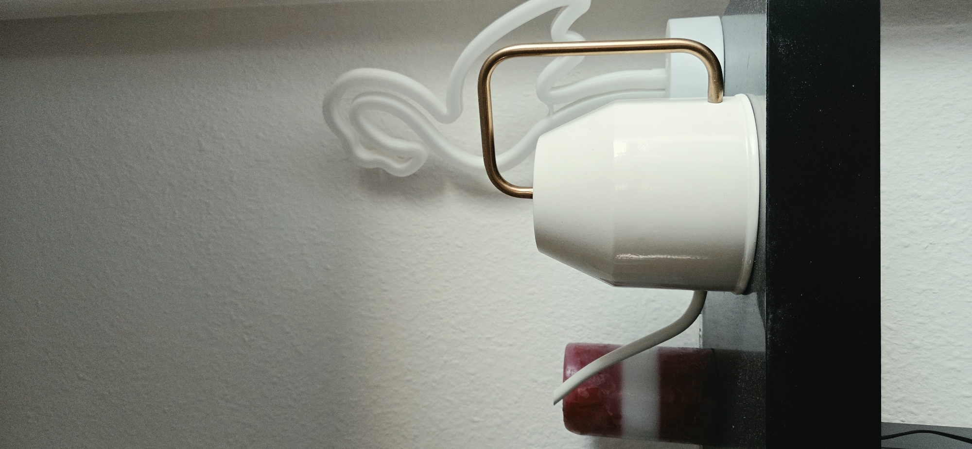 This image shows a still life arrangement on a black shelf against a textured white wall. On the left, there is a cylindrical candle with red and white stripes. To its right, a white watering can with a brass handle is centrally placed. In the background, a white neon flamingo light fixture is visible, creating a shadow on the wall. The scene is lit from the front, casting subtle shadows. The location is unknown.