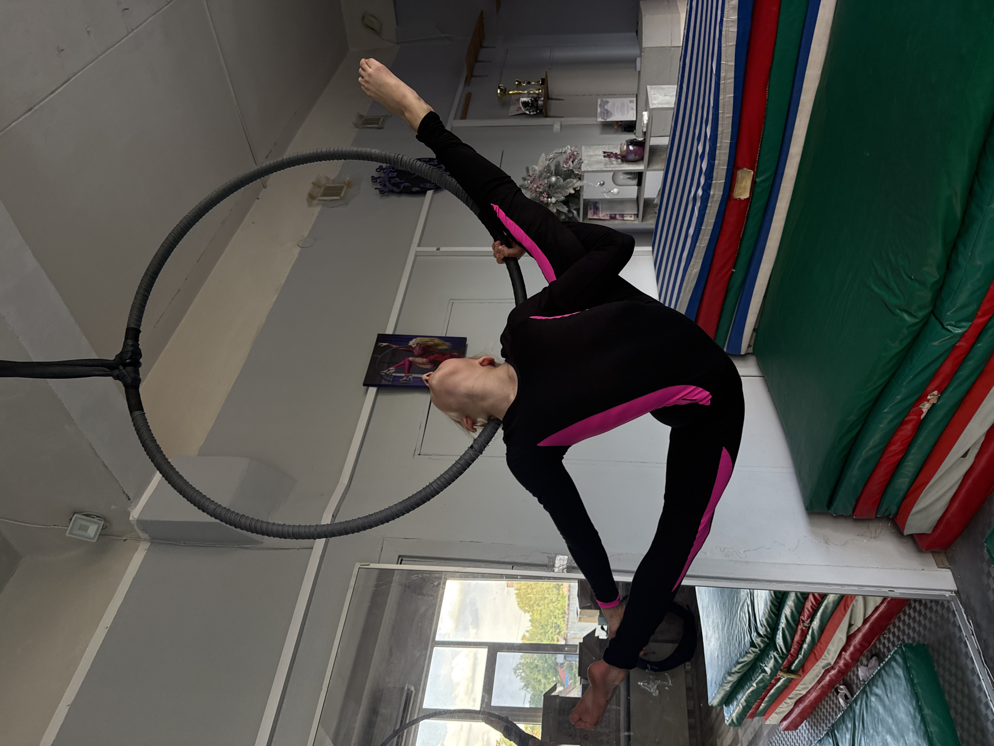 A young person in a black and pink full-body leotard performs an aerial hoop routine in a room with a high ceiling and light-colored walls. The hoop is suspended from the ceiling by a dark, ribbed cable. The person is positioned with one leg extended upwards and bent at the knee, while the other leg is extended horizontally. Their head is tilted back. In the background, a large mirror reflects a window showing trees and sky, as well as stacks of colorful mats. One set of mats is blue and white striped on top, followed by red and white, then green. Another stack of green mats is visible to the left. Shelving units with decorative items and trophies are present on the right side of the room. The lighting appears to be a mix of overhead and natural light. The specific location in Dnipro, Ukraine is not discernible from the image.