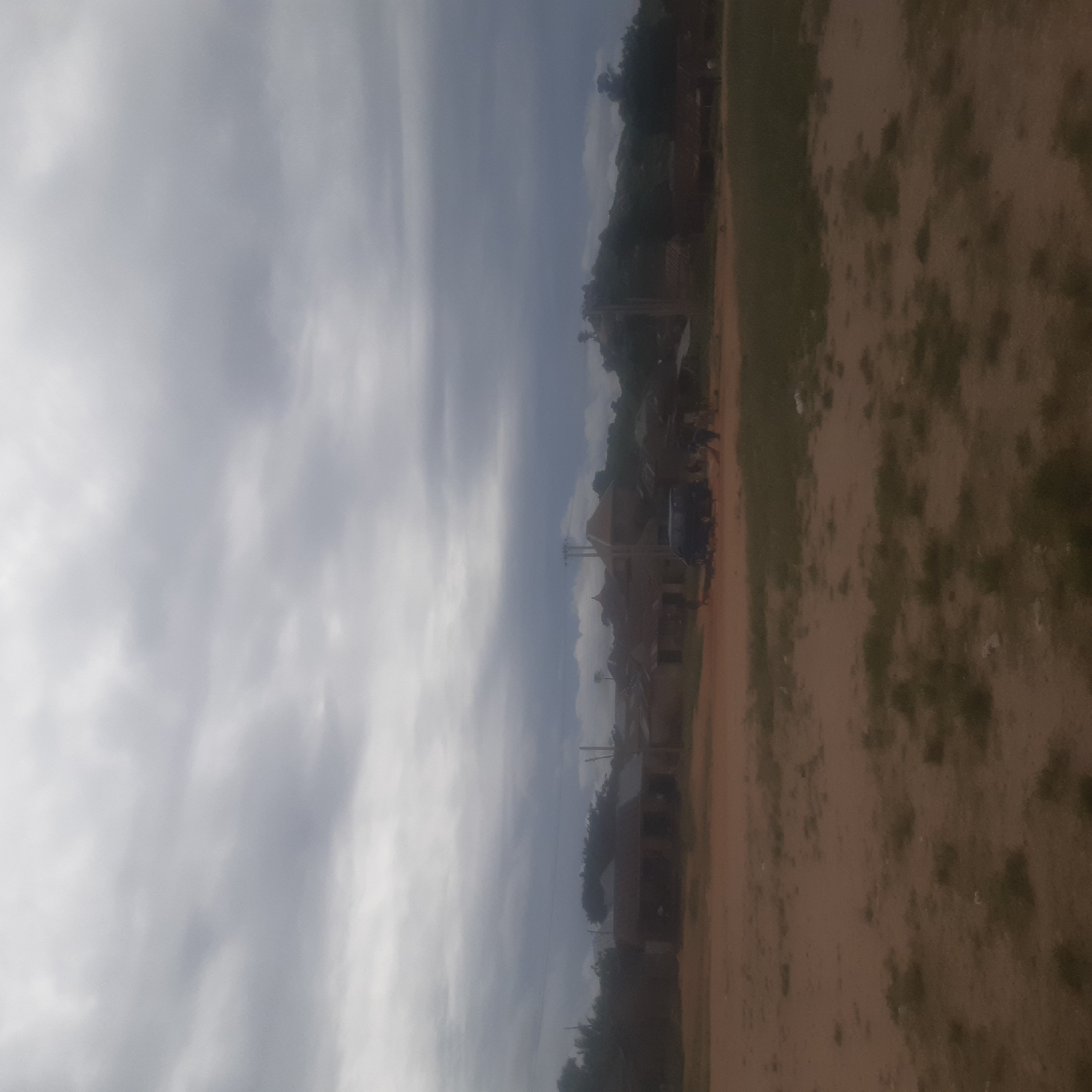The image depicts a rural or semi-urban scene with a dark, overcast sky. In the foreground, there is a dirt path with sparse green vegetation. In the middle ground, a cluster of buildings with corrugated iron roofs are visible. Some of these structures appear to be residential or commercial. A dark colored van is parked near the buildings, and several people are standing around it. The overall atmosphere is subdued due to the cloudy weather. The scene could be located in a village or outskirts of a town in Jos, Nigeria, given the architectural style of the buildings and the surrounding landscape.