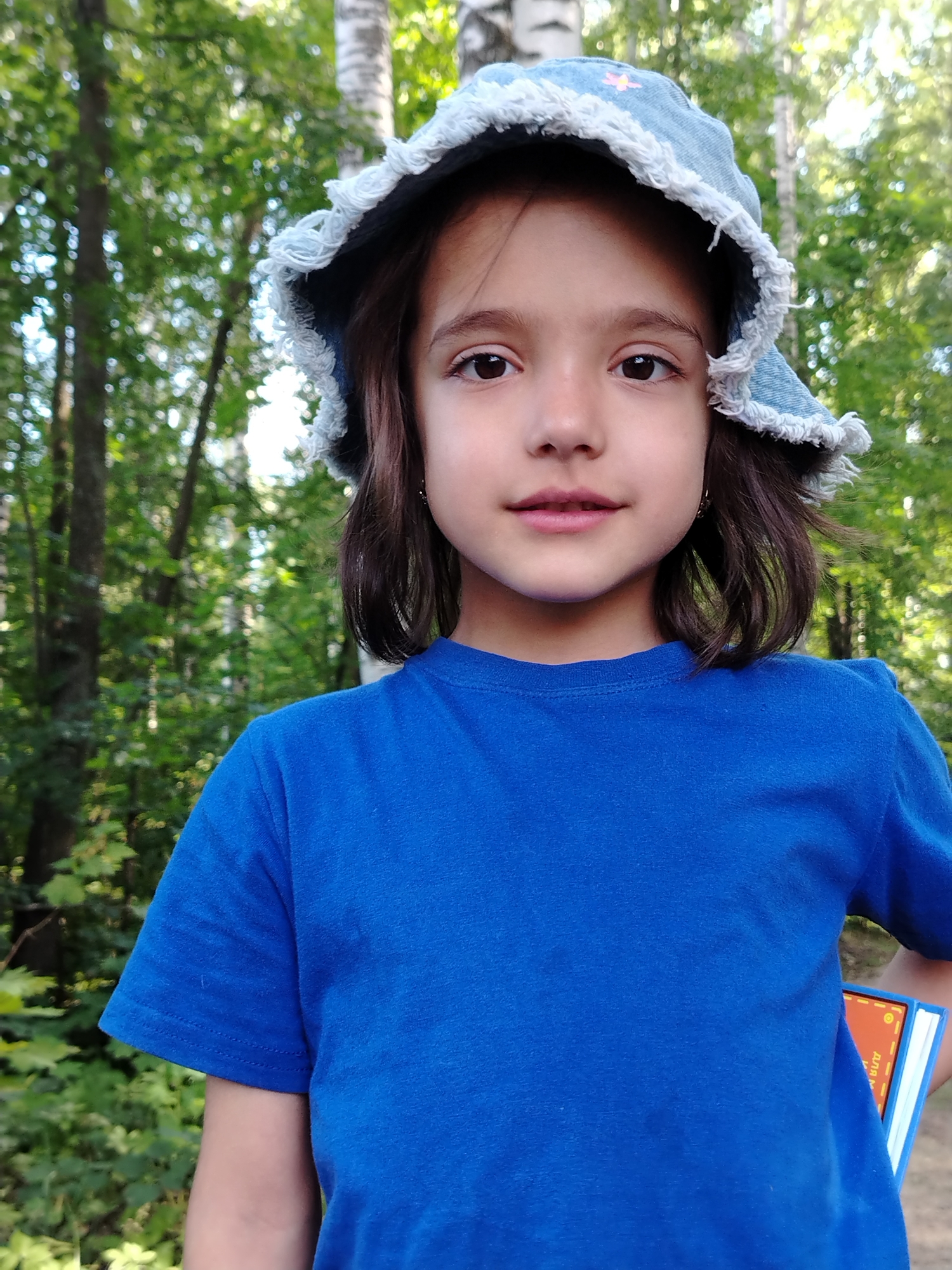 A young girl wearing a blue t-shirt and a denim bucket hat is photographed outdoors. The hat has a frayed edge and a small pink flower detail on the front. Her hair is dark brown and medium length. She is holding a blue book with an orange cover on her right side, partially obscured by her t-shirt. The background is blurred and shows a wooded area with trees and green foliage. The lighting suggests it is daytime. The photograph appears to be taken in Noviy, Russia, indicated by the text on the book.