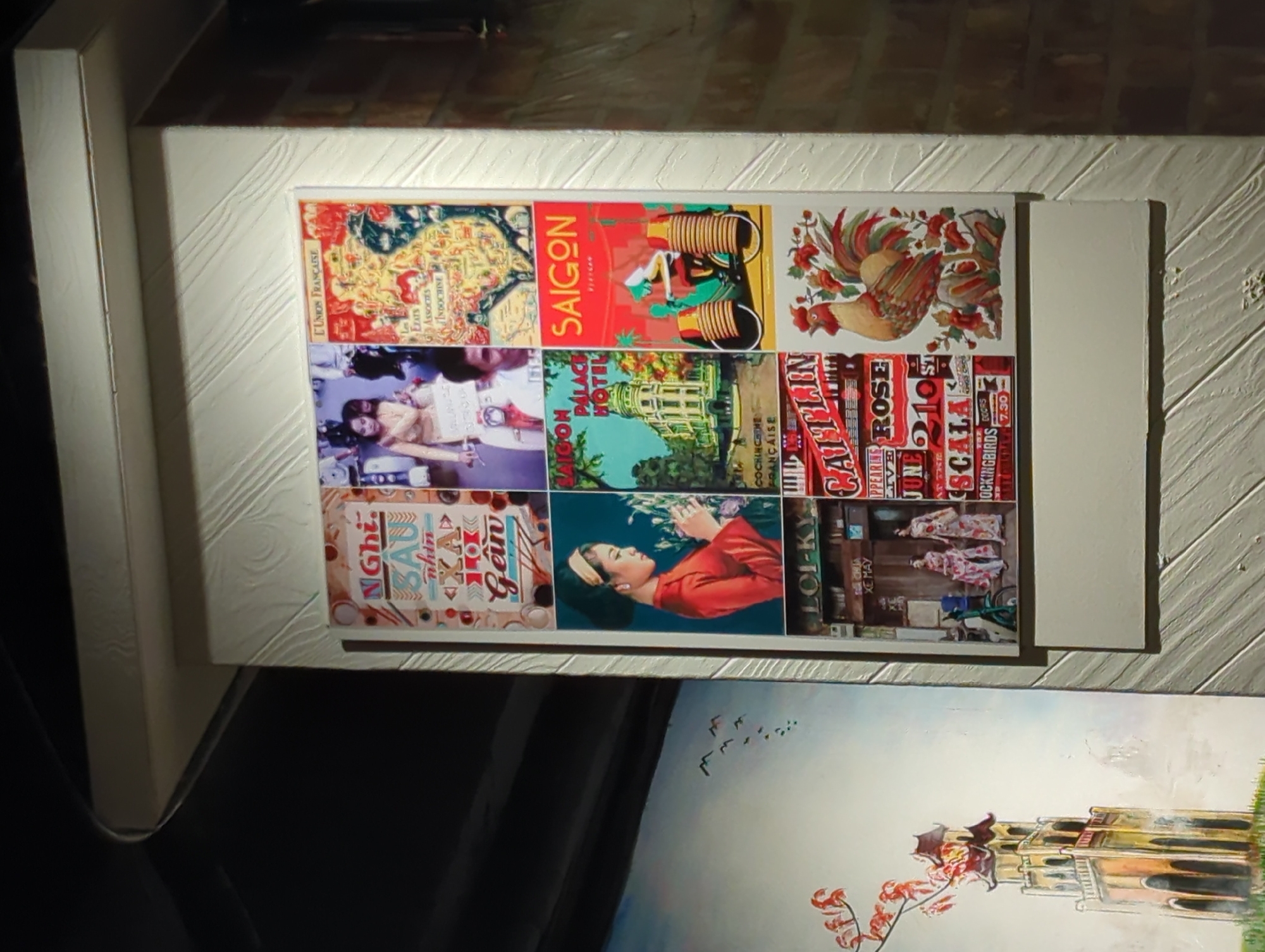 The image displays a collage of vintage-style posters mounted on a textured, cream-colored wall, which is positioned in a corner of what appears to be an indoor space with low lighting. The posters feature colorful illustrations and text, with a Vietnamese theme. One poster depicts a woman in traditional Vietnamese attire holding flowers, another shows a map of "L'UNION FRANCAISE," and a third features a rooster amidst flowers. Other posters include architectural scenes and stylized text. To the left of the collage, a separate large painting is partially visible, showing a pagoda-like structure with red flowers on a branch. The wall's texture is a prominent detail, with a diagonal, wood-like pattern.