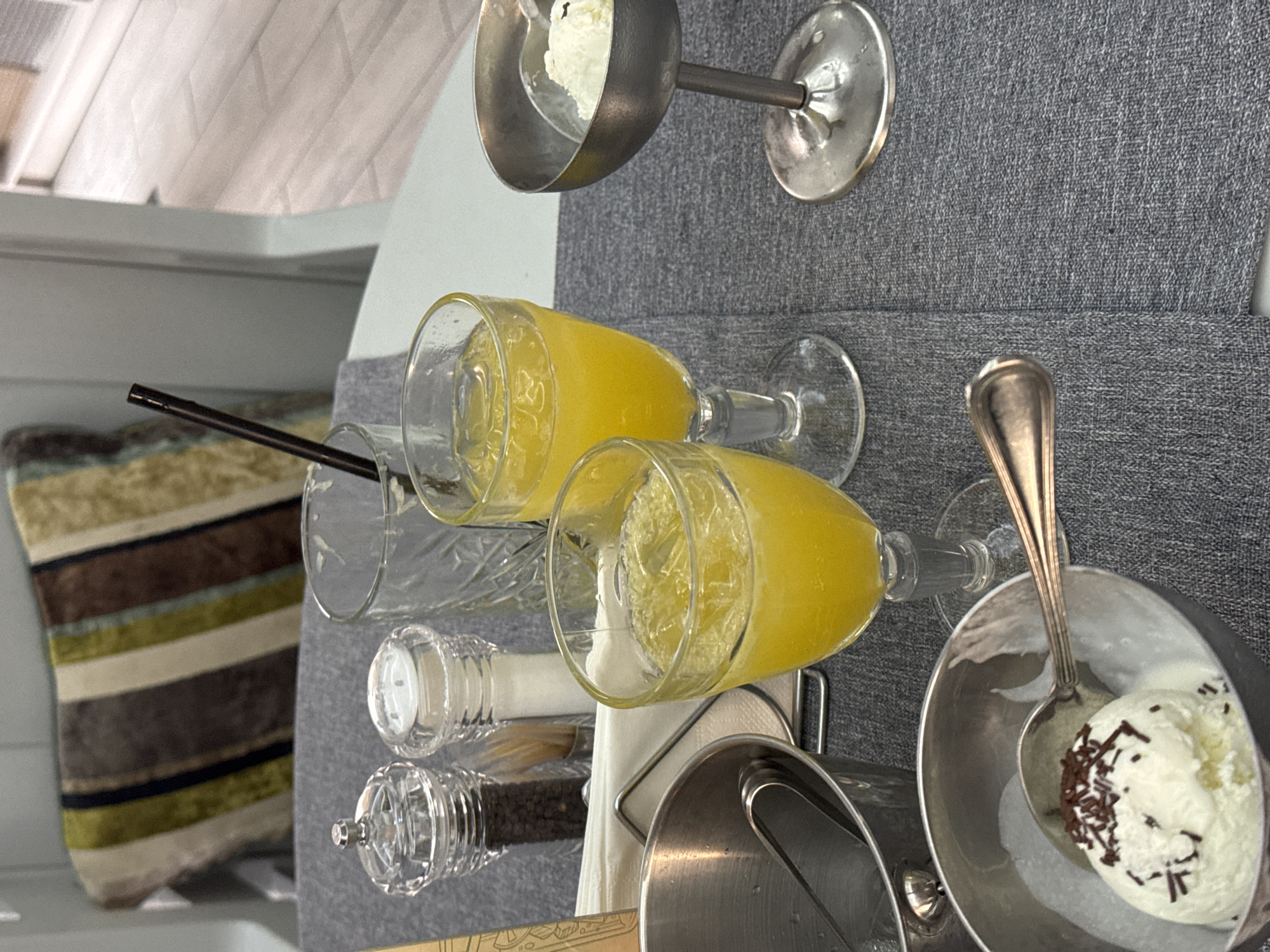 A close-up, eye-level shot captures a table setting with two glasses of orange juice filled with ice, a silver ice bucket with tongs, a silver goblet containing a scoop of vanilla ice cream topped with chocolate sprinkles, and another silver serving dish with ice cream. Also visible are salt and pepper shakers, a glass with a straw, and a white napkin. The table is covered with a grey textured tablecloth, and a striped cushion is visible in the background. The scene appears to be indoors. This image was taken in Sovkhoz imeni Lenina, Russia.