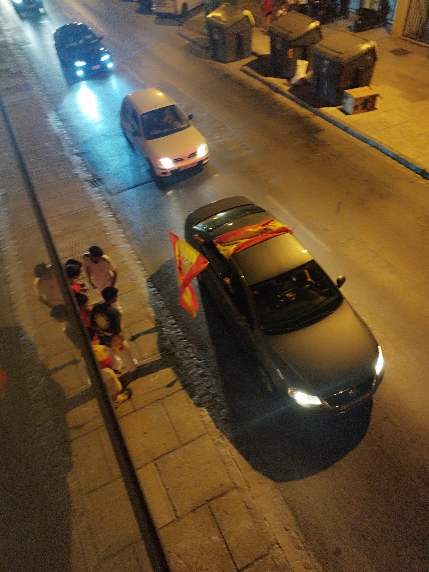 In Jerez, Spain, a dark-colored sedan with its headlights illuminated travels down a street at night. A Spanish flag is draped across the roof of the car. Several individuals are standing on a sidewalk to the left of the car. One of the individuals appears to be holding a flag as well. The sidewalk is paved with light-colored square tiles. In the background, several large, dark trash receptacles are visible, along with a small, wooden structure.