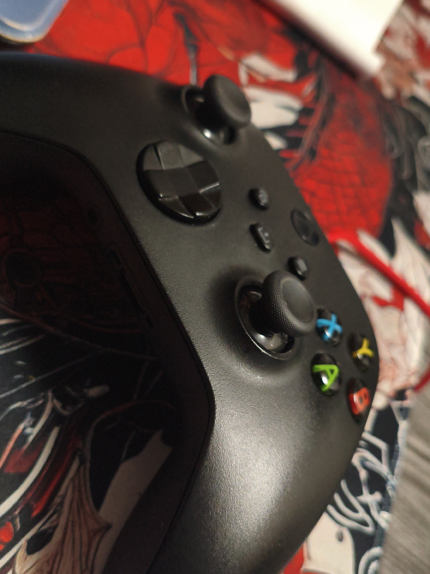 The image shows a close-up, high-angle shot of a black Xbox controller resting on a red and white patterned surface, possibly a mat or cloth. The controller is positioned diagonally across the frame, with the analog sticks and face buttons clearly visible. The face buttons are colored green (A), blue (X), yellow (Y), and red (B), consistent with an Xbox controller. The surface beneath the controller features a busy, graphic design with abstract shapes and possibly figures, predominantly in red and white. A red wire is visible extending from the upper right corner of the frame. The lighting appears to be natural or from an overhead source, casting subtle shadows on the controller's surface. There are no people or animals visible in the image. The image was likely taken indoors, given the focus on the controller and the textured surface it rests on. There is no information in the image to suggest a location in Vladivostok, Russia.