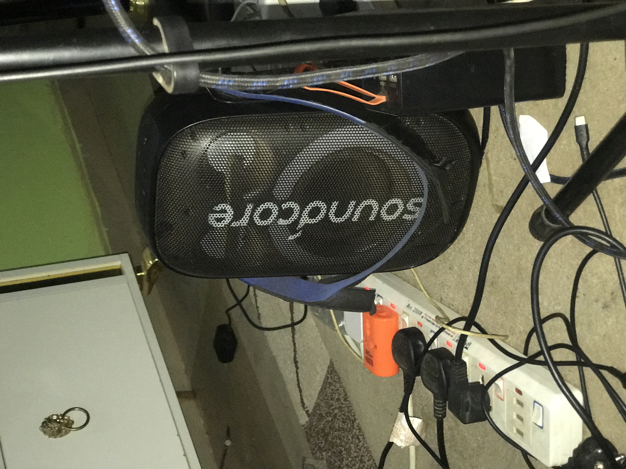 The image displays a cluttered arrangement of electronic equipment and cables, likely situated under a desk or a similar workstation. A prominent black Anker Soundcore portable speaker lies horizontally in the center, its mesh grille showing the white "soundcore" logo.
Numerous electrical cables, primarily black and blue, are entangled around the speaker and connect to a white power strip located on the floor or ground. The power strip features multiple outlets with individual switches and prominently displays the text "Max. 3500W." Several power plugs are inserted into the strip, including distinctive black three-pin rectangular plugs (Type G, common in Nigeria) and an orange plug. A white USB-C cable end is also visible among the wiring.
Above the speaker and cables, black metal bars or structures suggest the framework of a desk or stand. To the left, a section of a green surface (possibly a wall or mat) borders a white furniture item, likely a drawer or cabinet, upon which a metallic key ring adorned with keys or charms rests. The floor beneath appears to be a beige or grey textured surface. The scene portrays a functional but somewhat untidy electronic setup.