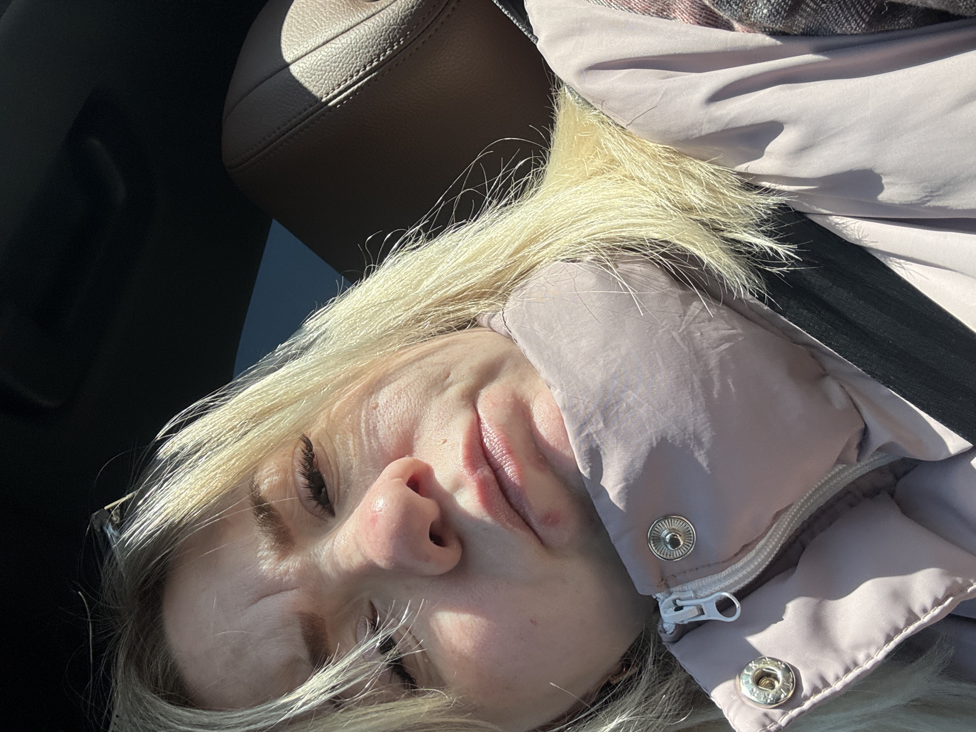 A selfie image depicts a fair-skinned blonde woman with her eyes closed. She is wearing a light pink puffer jacket and a dark-colored seatbelt is visible across her chest. Part of a brown leather car seat is visible in the upper left corner. The location and city details cannot be determined from the image.