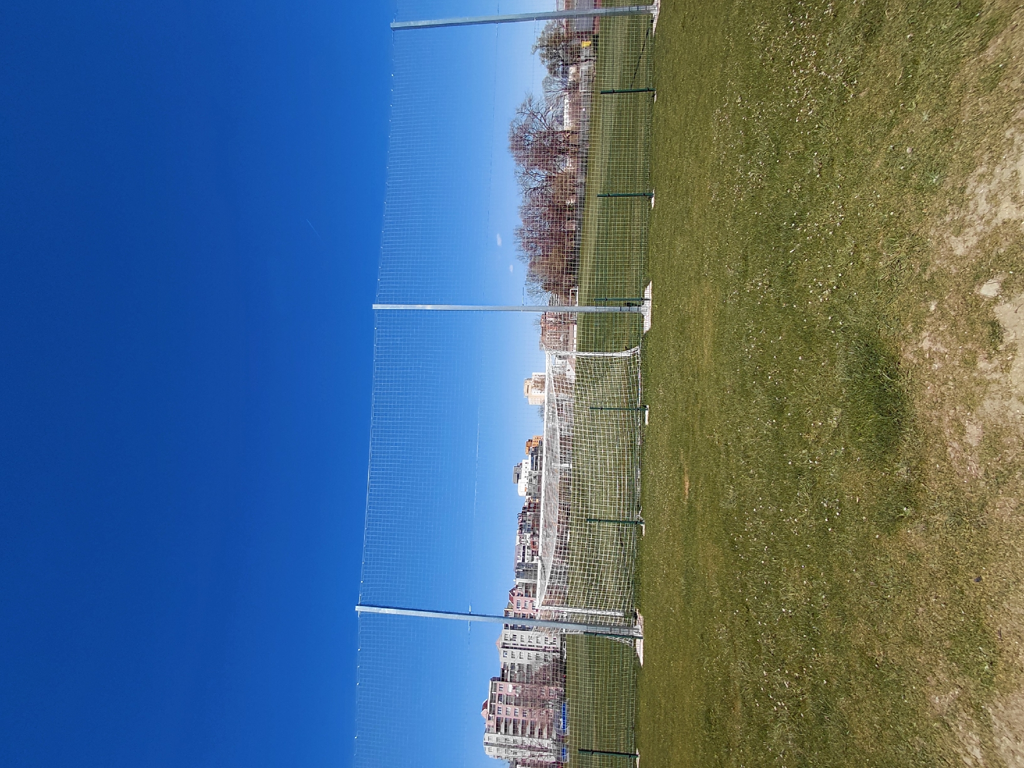 The image shows a soccer field in Sofia, Bulgaria, on a sunny day. The sky is a clear, bright blue. The field is green and bordered by a fence. In the background, buildings can be seen. Two soccer goals are visible on the field. There are no people visible in the image. The scene is well-lit.